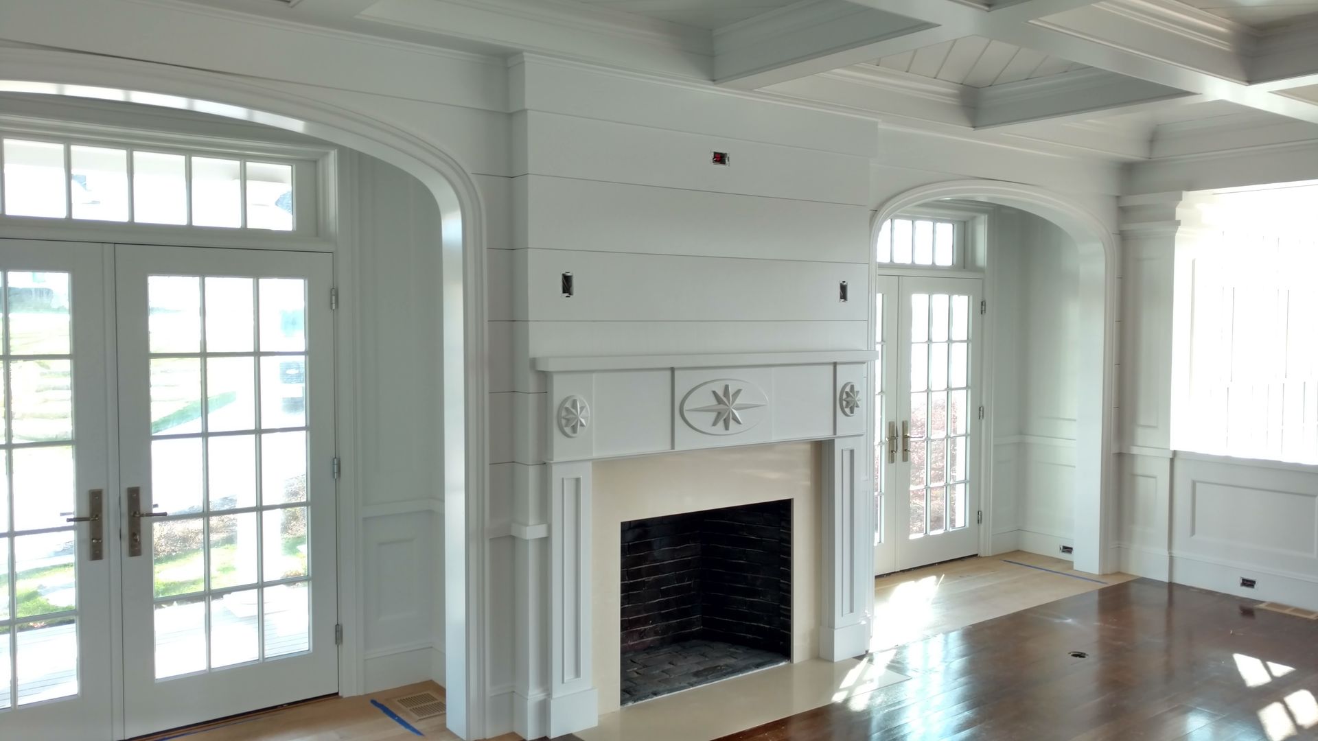 An interior room with white paneled walls, a central fireplace, coffered ceilings, and white-framed French doors.