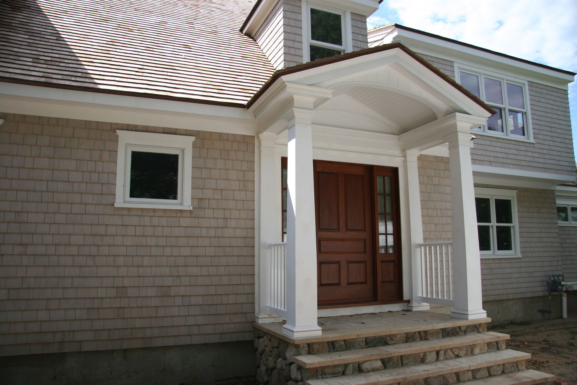 A tan shingled house exterior with a front porch, white columns, a wooden door, and stone-trimmed steps.
