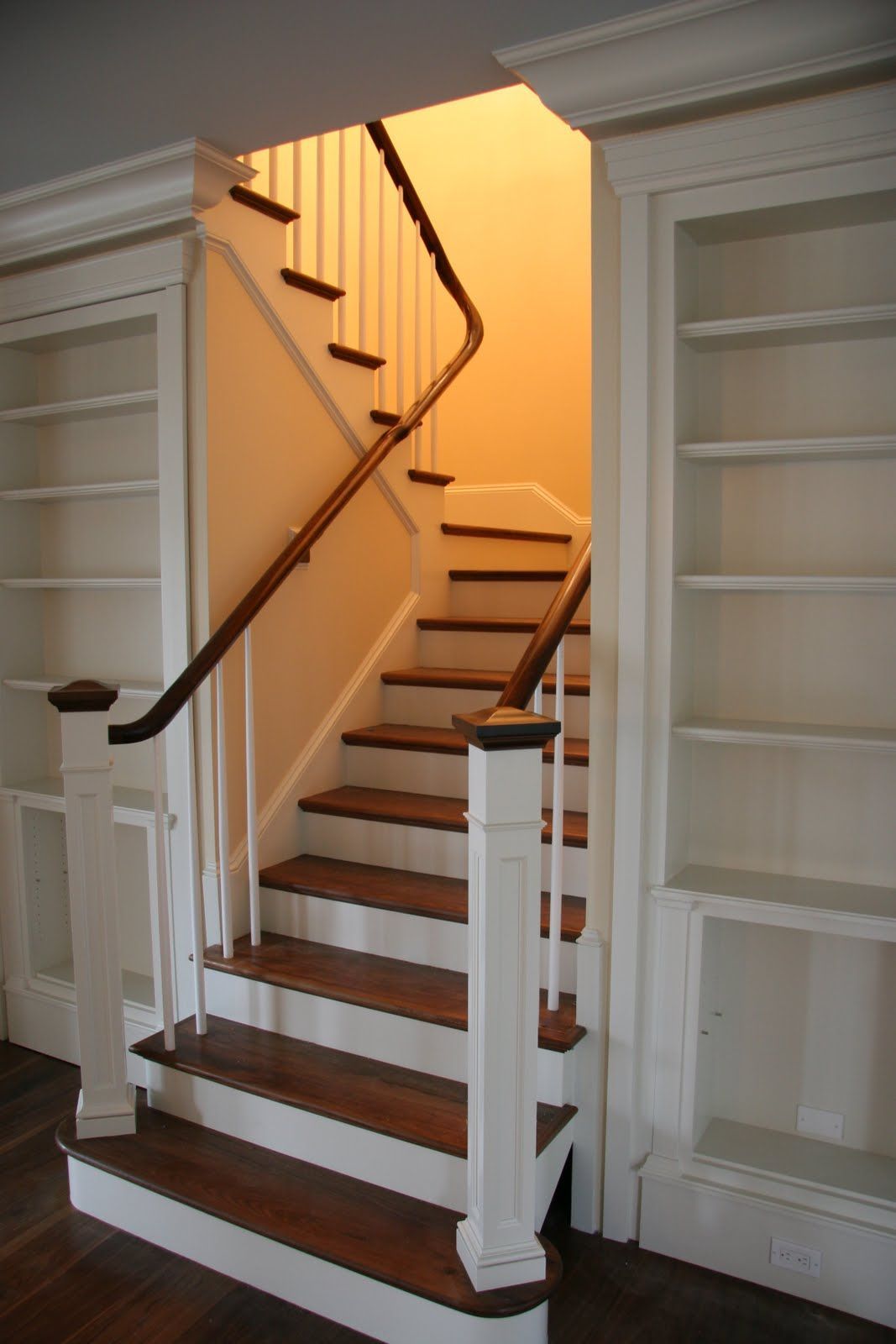 A staircase with dark wood treads and white risers, flanked by two built-in white bookshelves, leading to a warm-lit area.