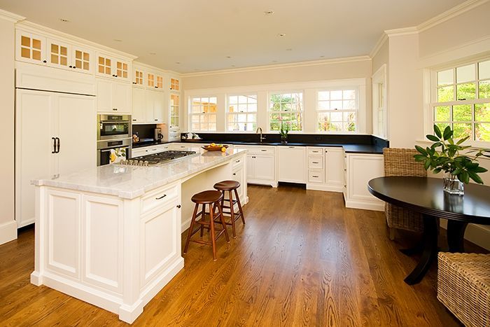 A bright kitchen with white cabinets, a large marble-topped island, wooden floors, and a small round black dining table.