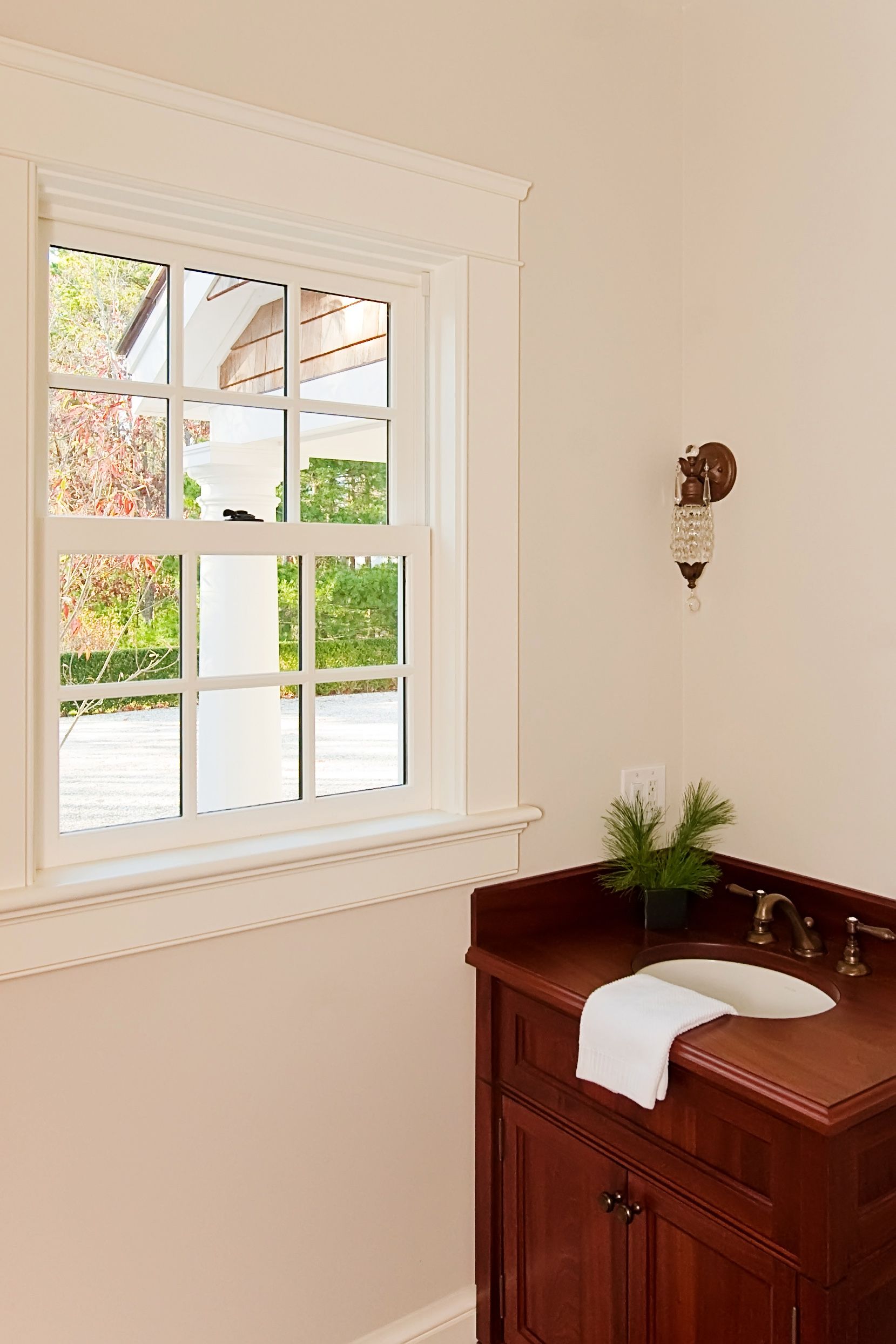 A wood vanity with a white towel and small plant sits beside a window with cream trim in a bright, neutral-toned room.