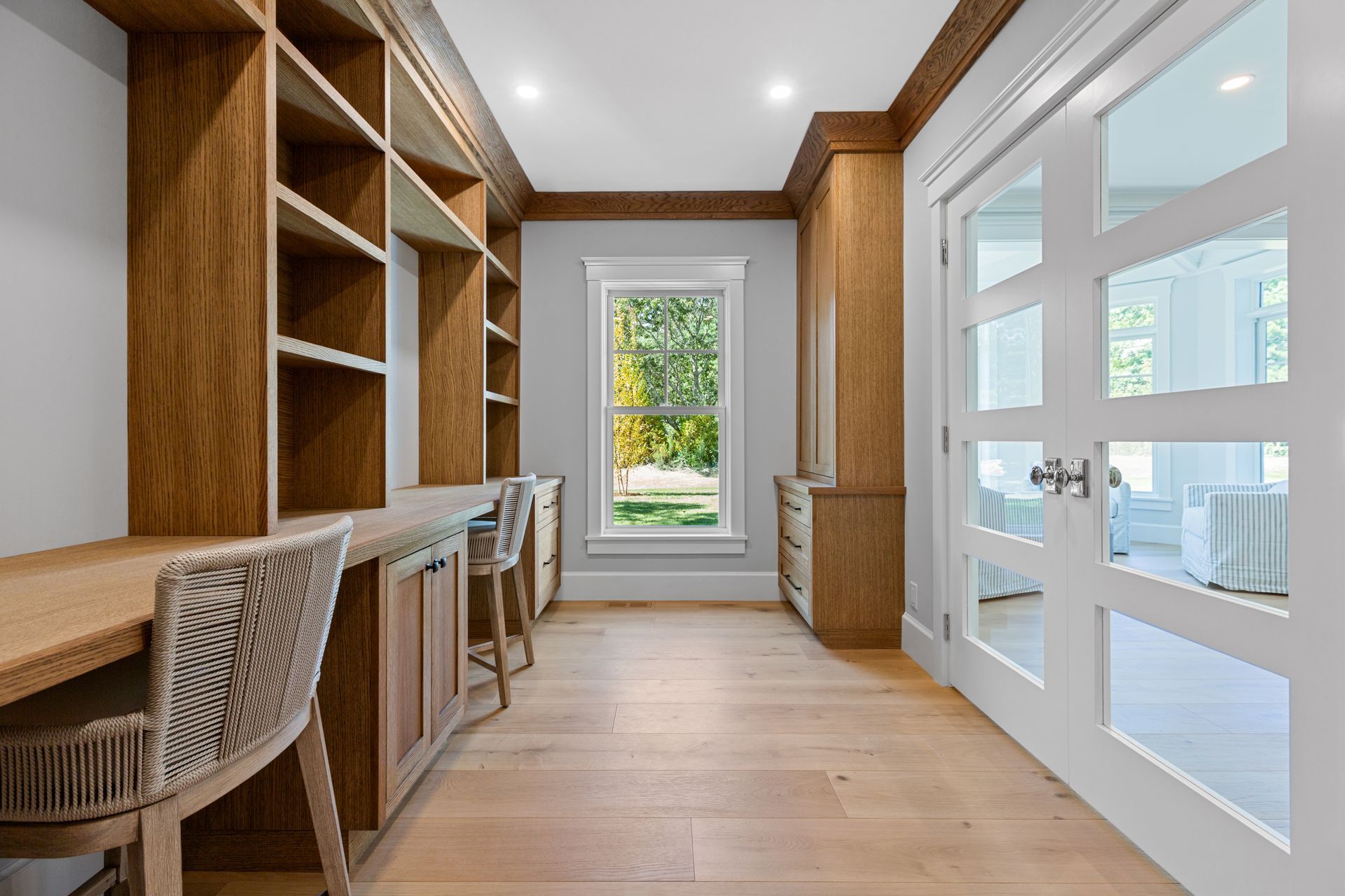 A home office features light wood floors, a built-in desk with wooden shelves, and white French doors leading to a sunroom.