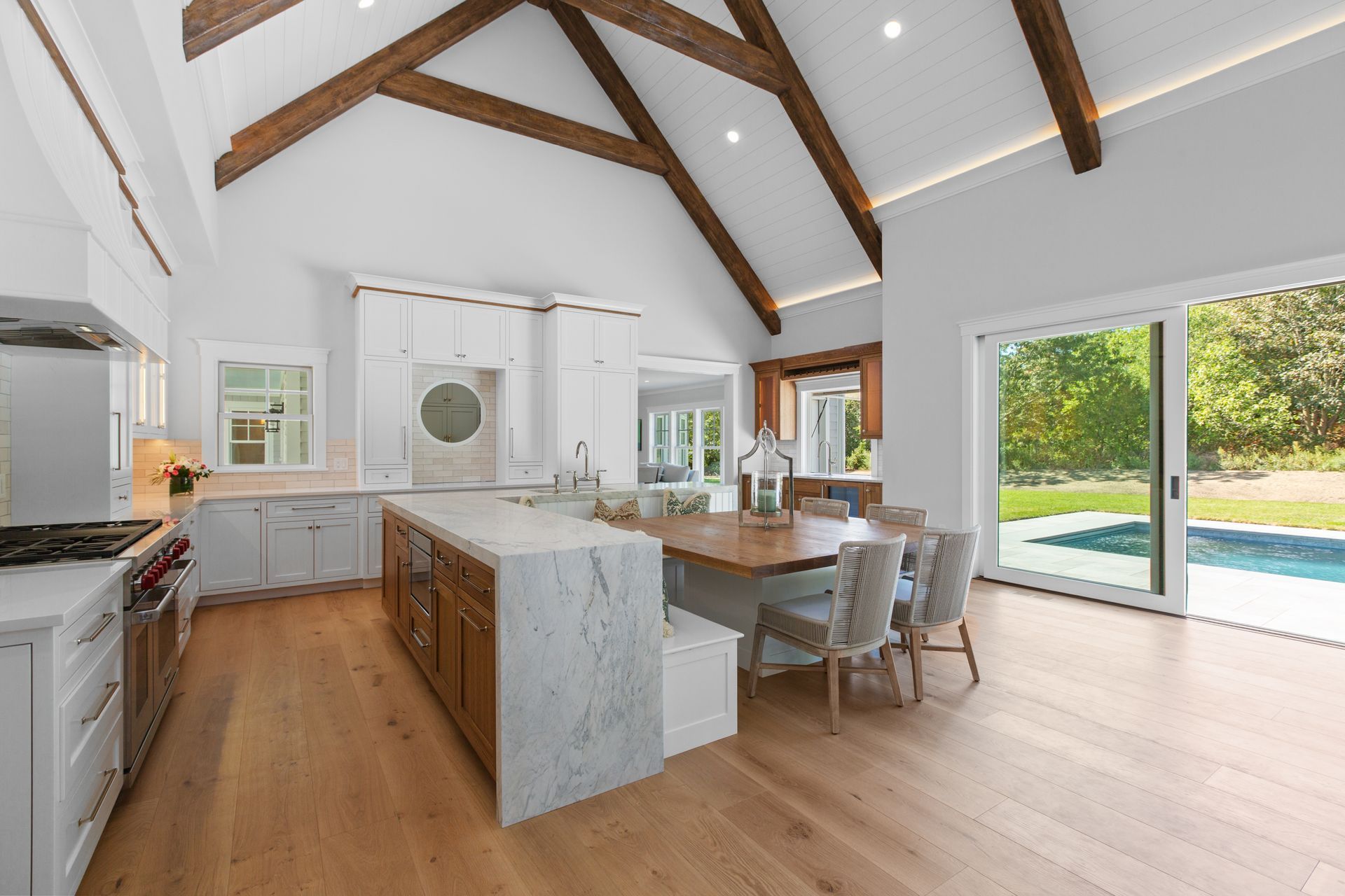 Bright, modern kitchen with white cabinetry, a marble-topped island, wooden dining table, and vaulted wood-beamed ceiling.