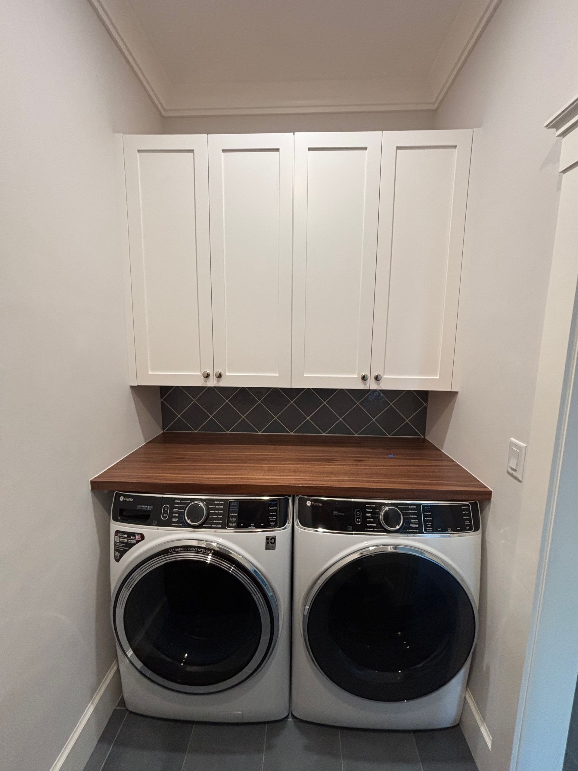 A laundry room featuring two white washing machines, a dark wood countertop, grey herringbone backsplash, and white cabinets.