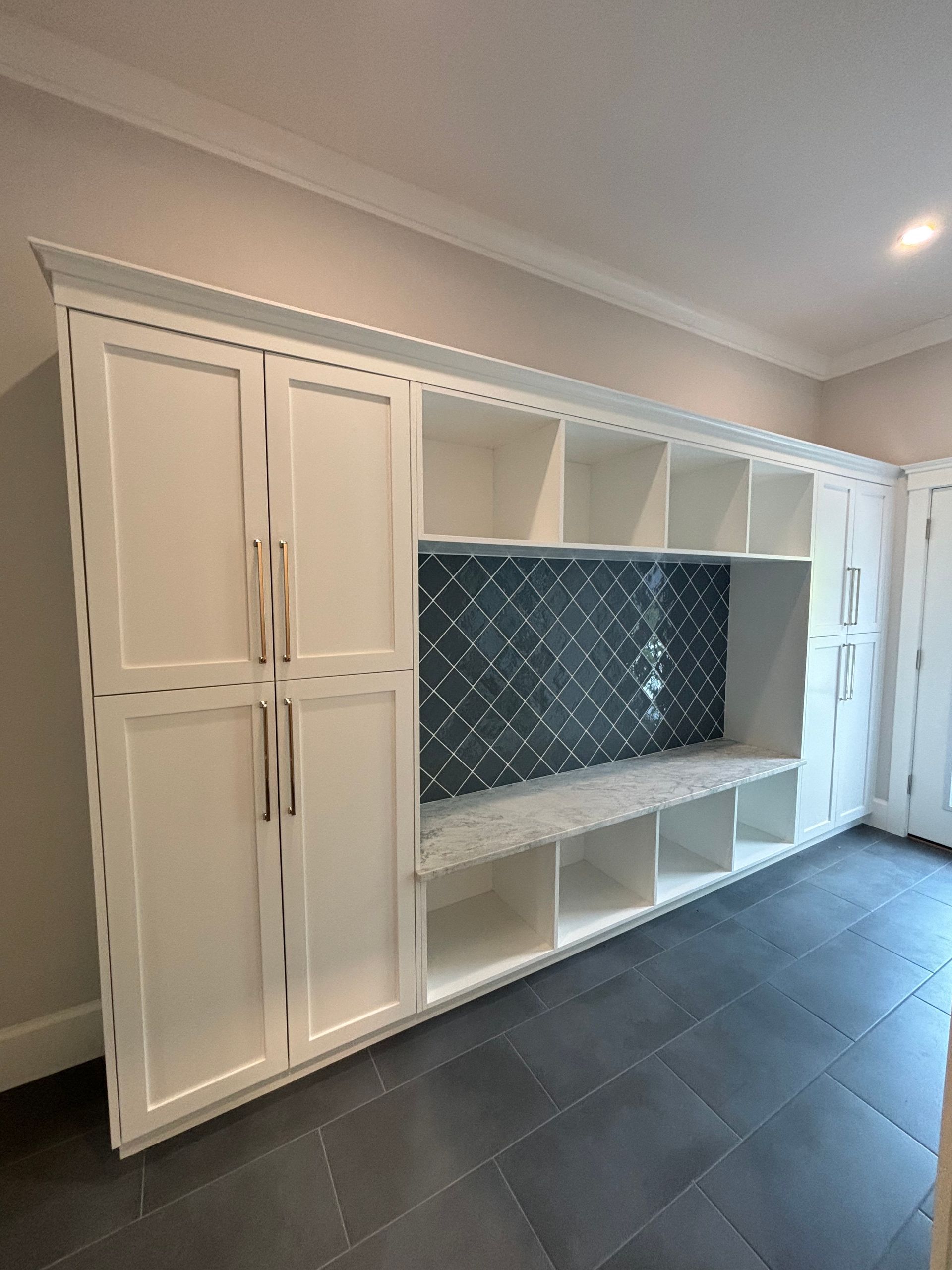 Custom white built-in mudroom cabinetry with a tiled backsplash, bench seating, and storage shelves on a grey tile floor.