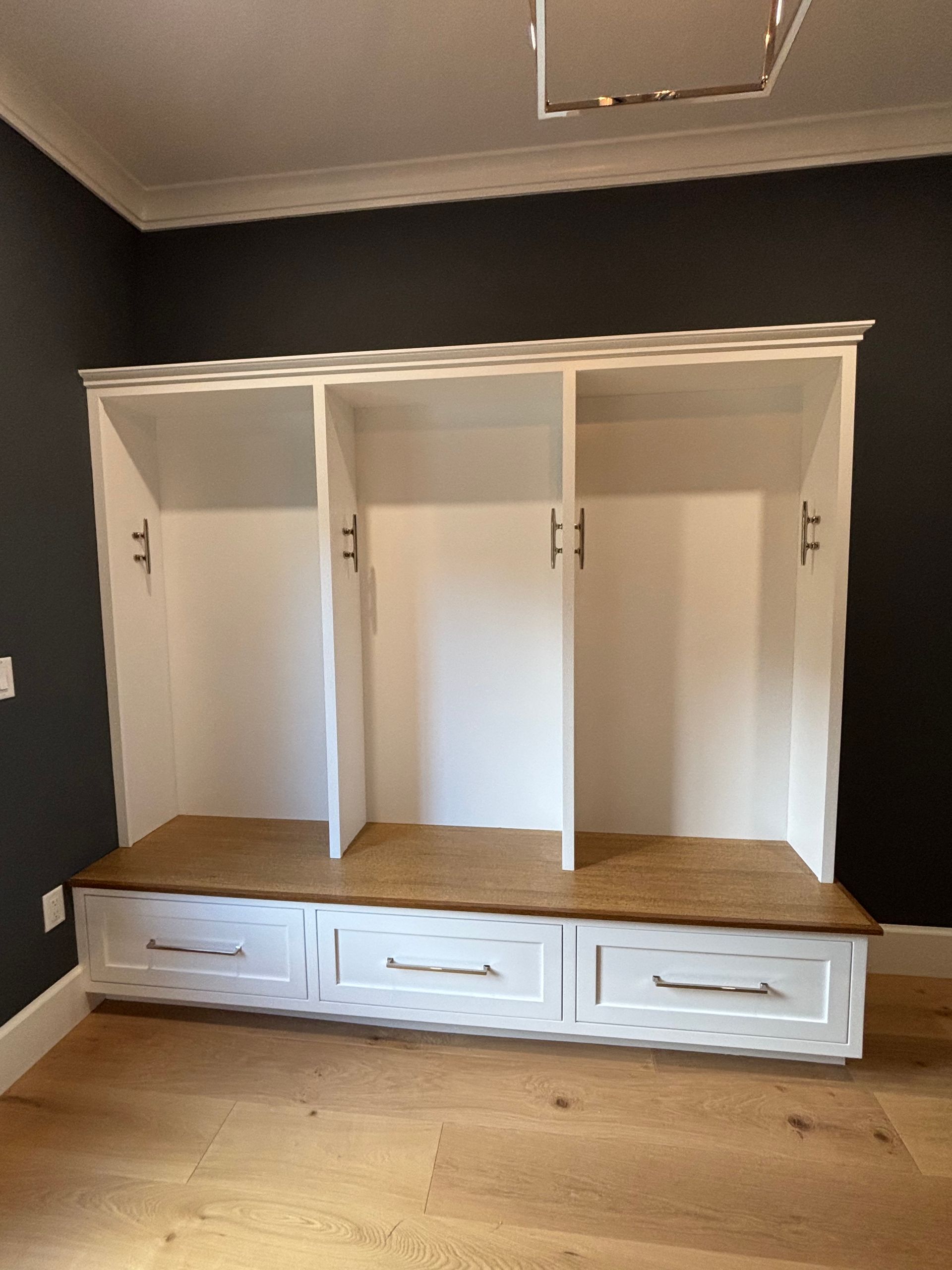 A three-section white mudroom bench with wooden seating, three lower drawers, and silver hardware against a dark wall.