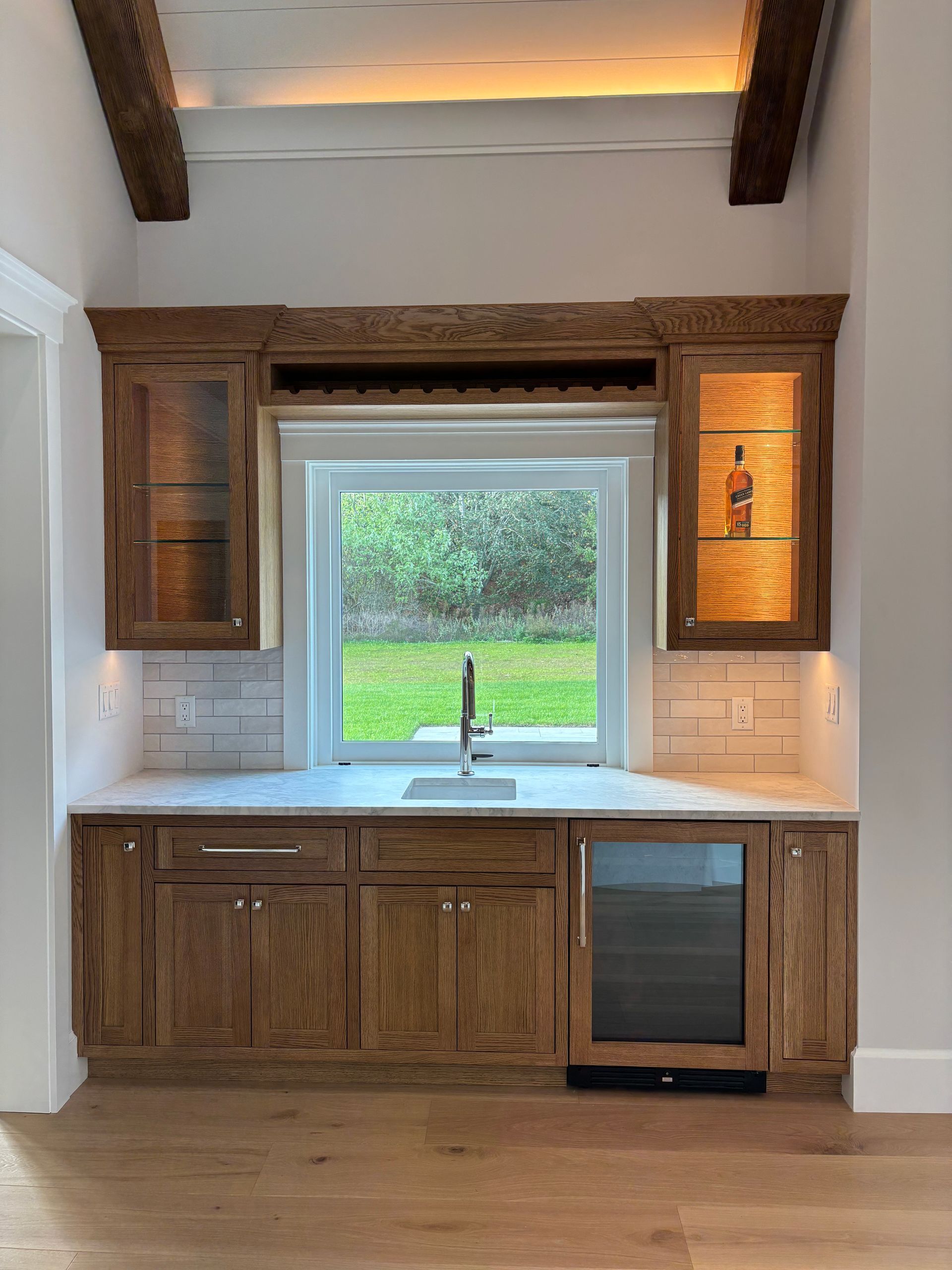 A wet bar with rustic oak cabinets, under-cabinet lighting, a built-in wine fridge, and a sink beneath a window.