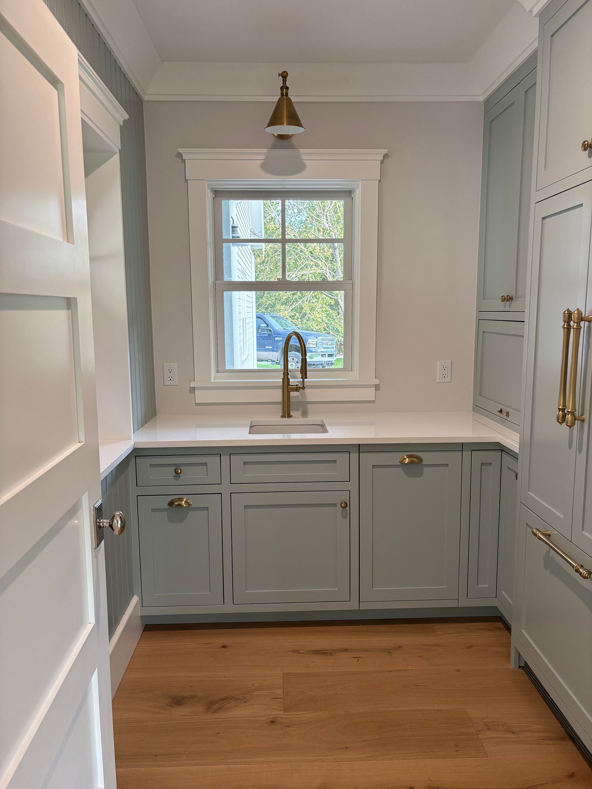 A small, light blue galley kitchen with white trim, a window over the sink, and light wood floors.