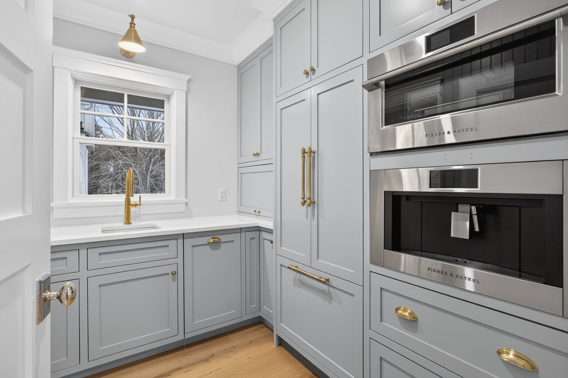 Light blue kitchen cabinetry with gold hardware, stainless steel appliances, and a window.