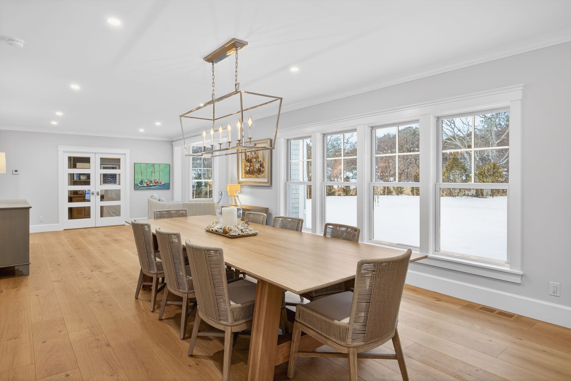 Dining room with light wood table, woven chairs, large windows, and a rectangular chandelier.