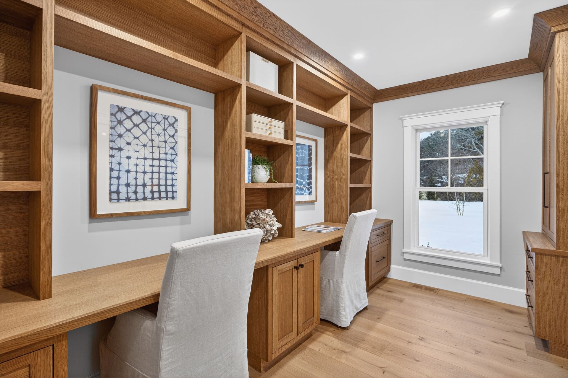 Home office with built-in wooden shelving, desks, and two white chairs. A window is in the background.