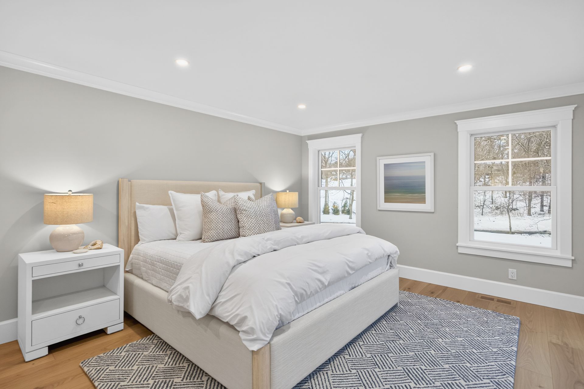 Bedroom with a light-colored bed, bedside table, rug, and windows with white trim. Gray walls.