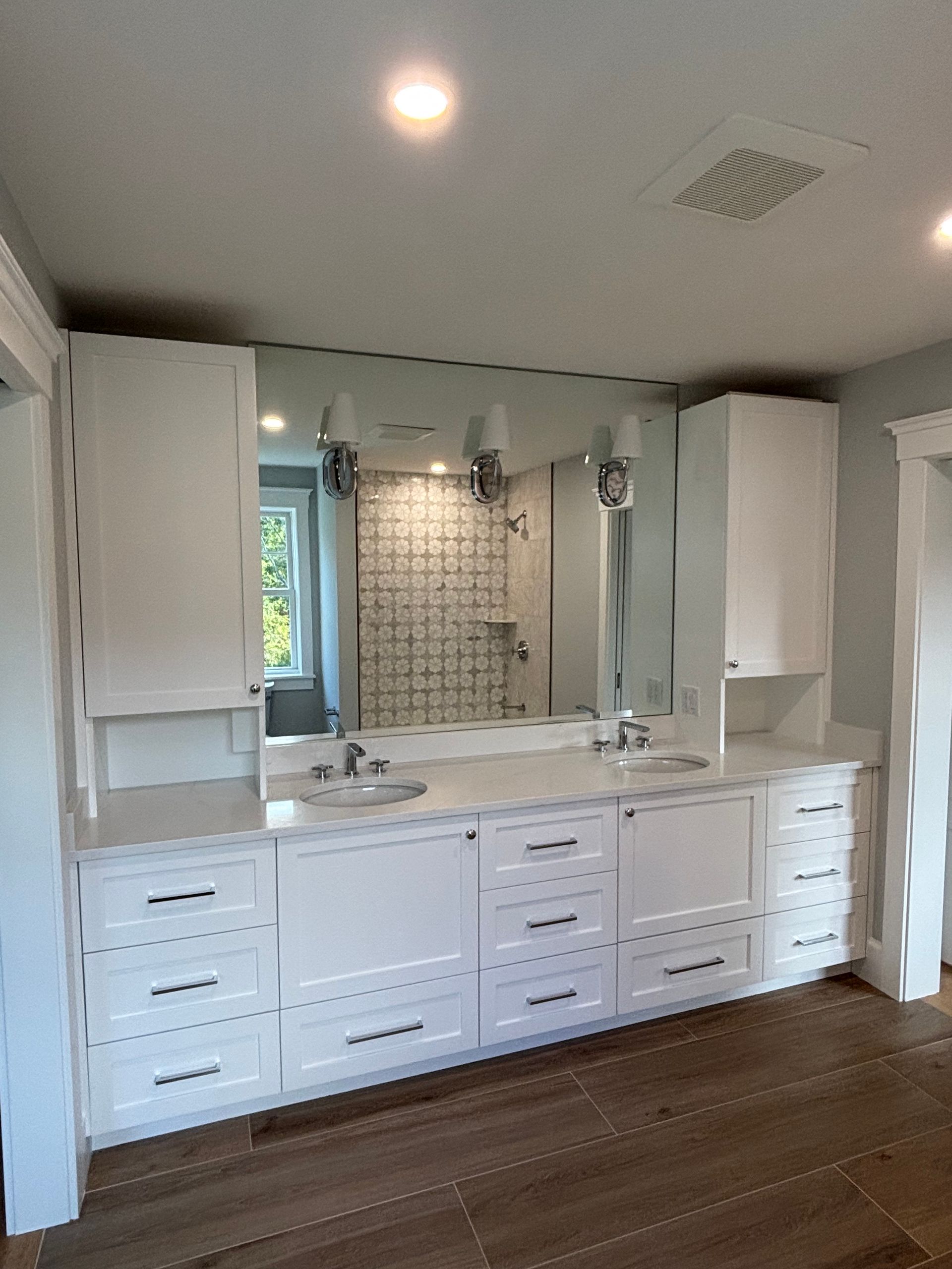 A white double vanity with cabinets, drawers, and sinks in a bathroom with wood-look tile floors and a large mirror.