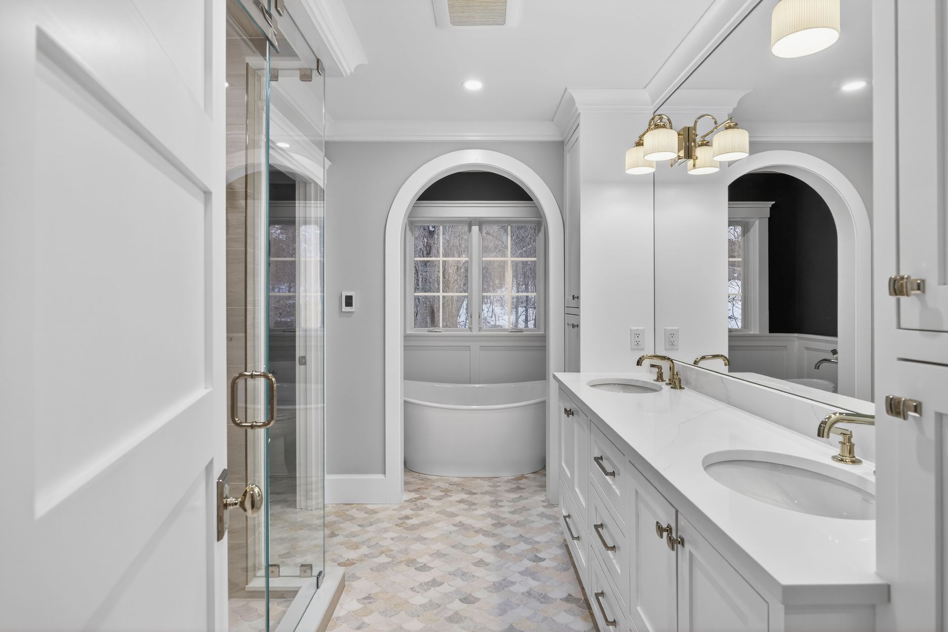 Elegant white bathroom with gray walls, glass shower, double sinks, and arched window.