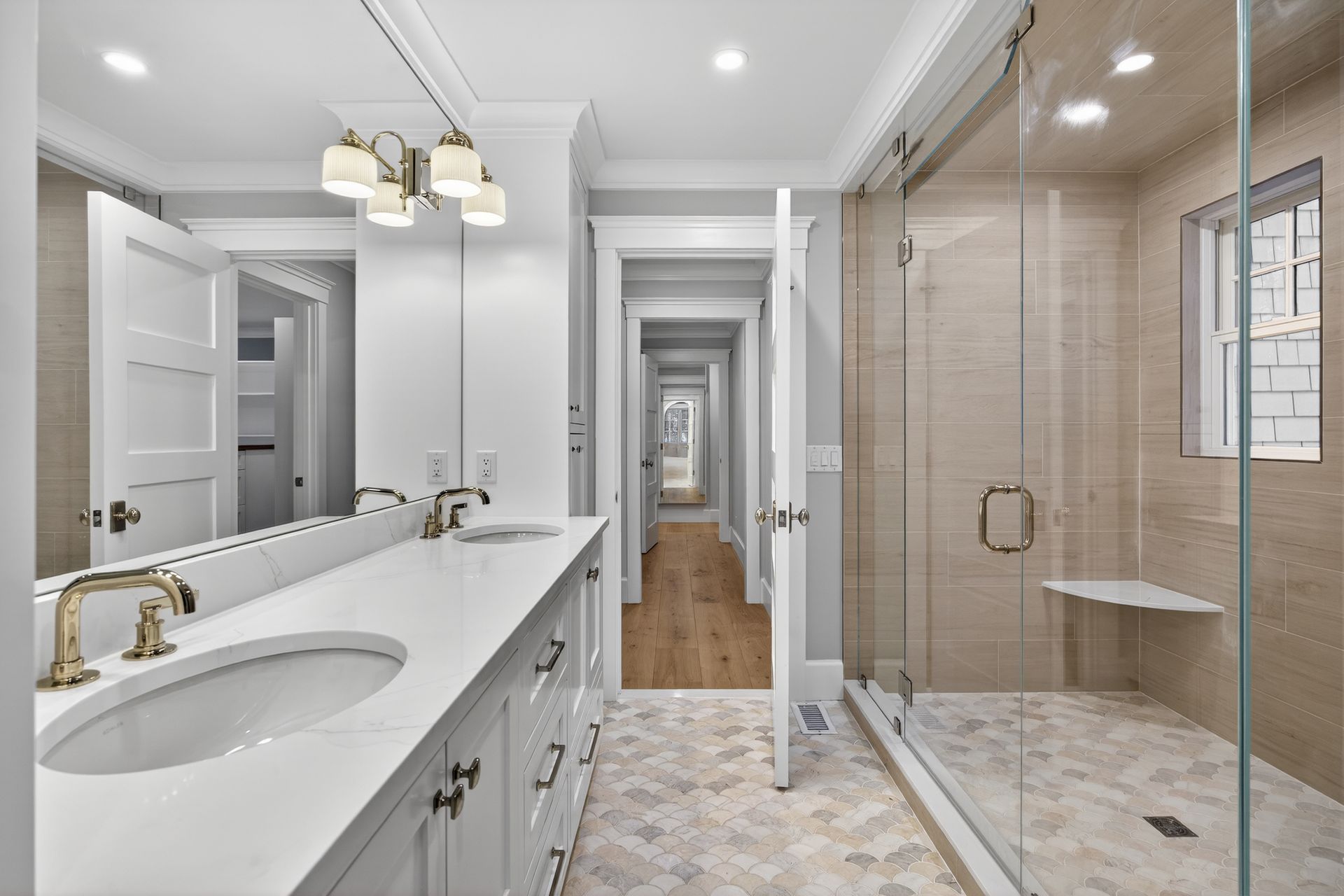 Bathroom with a long vanity, glass shower, and hallway. Light colors, gold fixtures, and tiled floor.