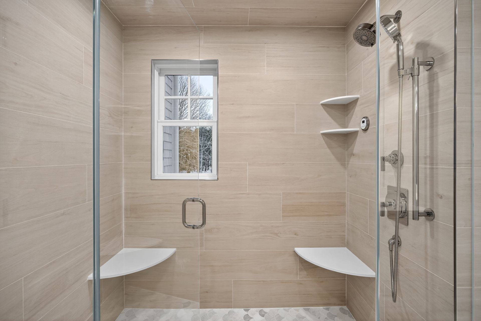 Modern shower with beige tiled walls, glass doors, and two built-in seats; a window is centered.