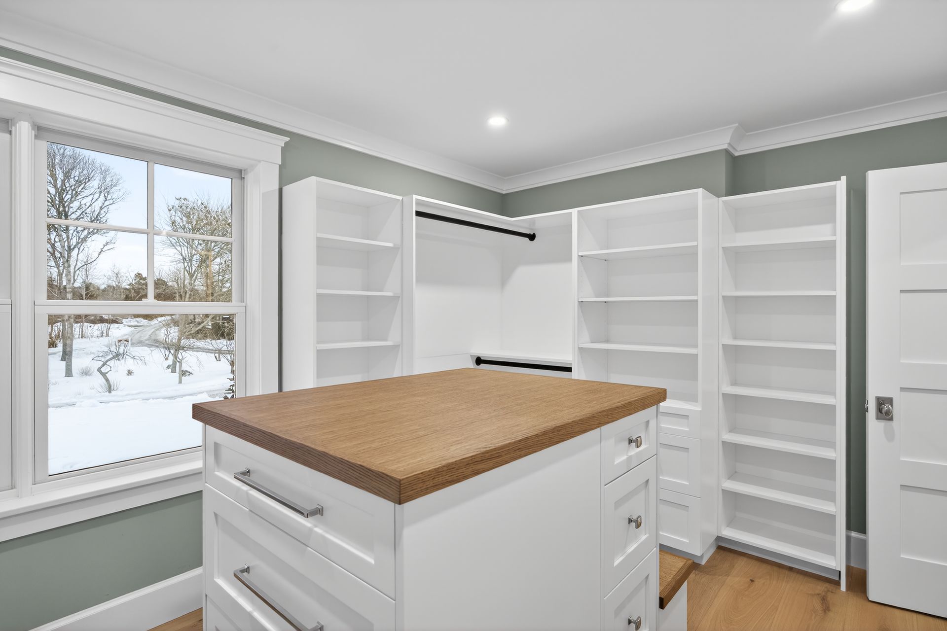 Walk-in closet with white shelves and drawers, wooden countertop, and window overlooking a snowy landscape.