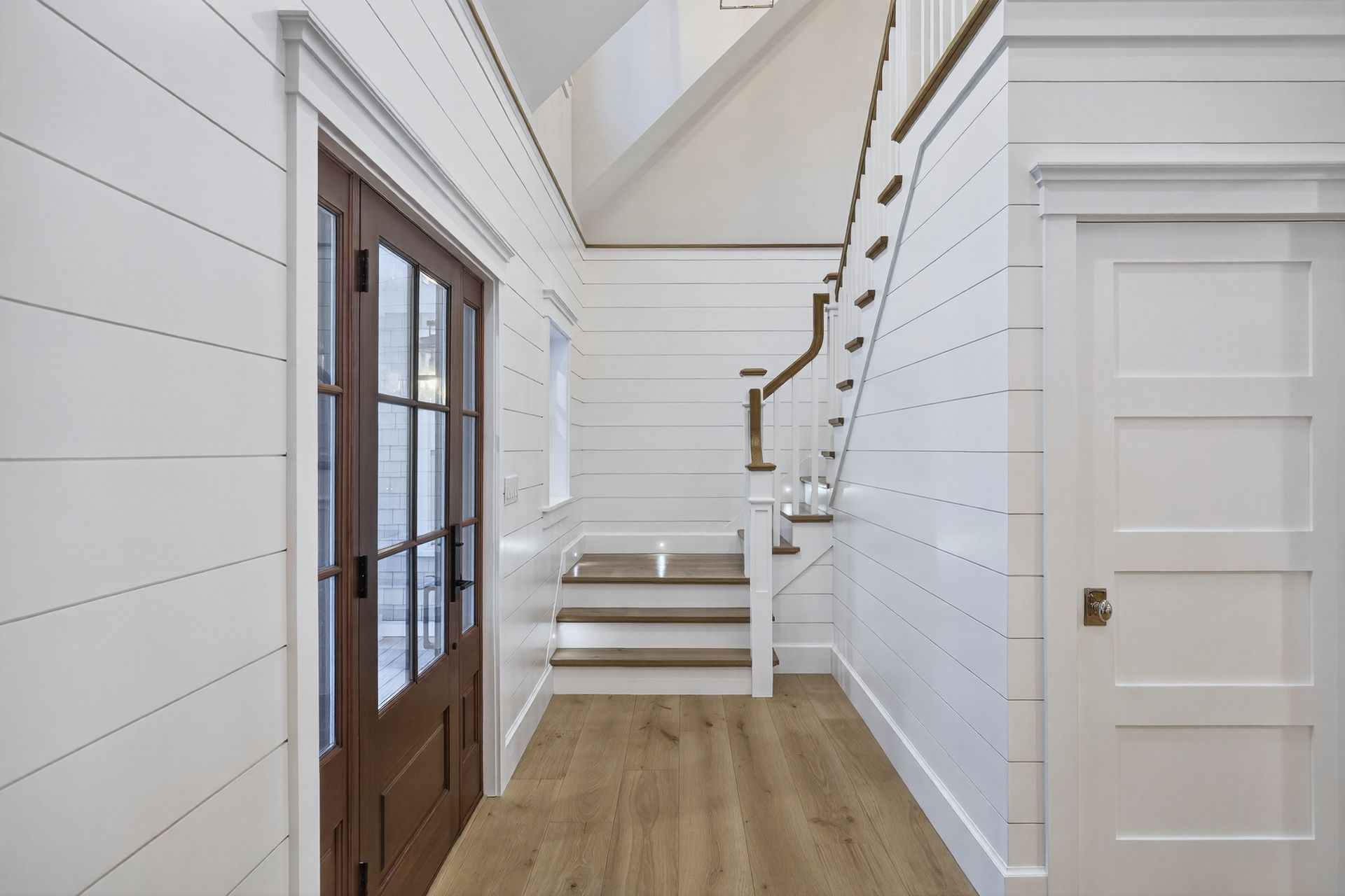 White paneled hallway with stairs, brown door, and white doors. Light wood flooring.