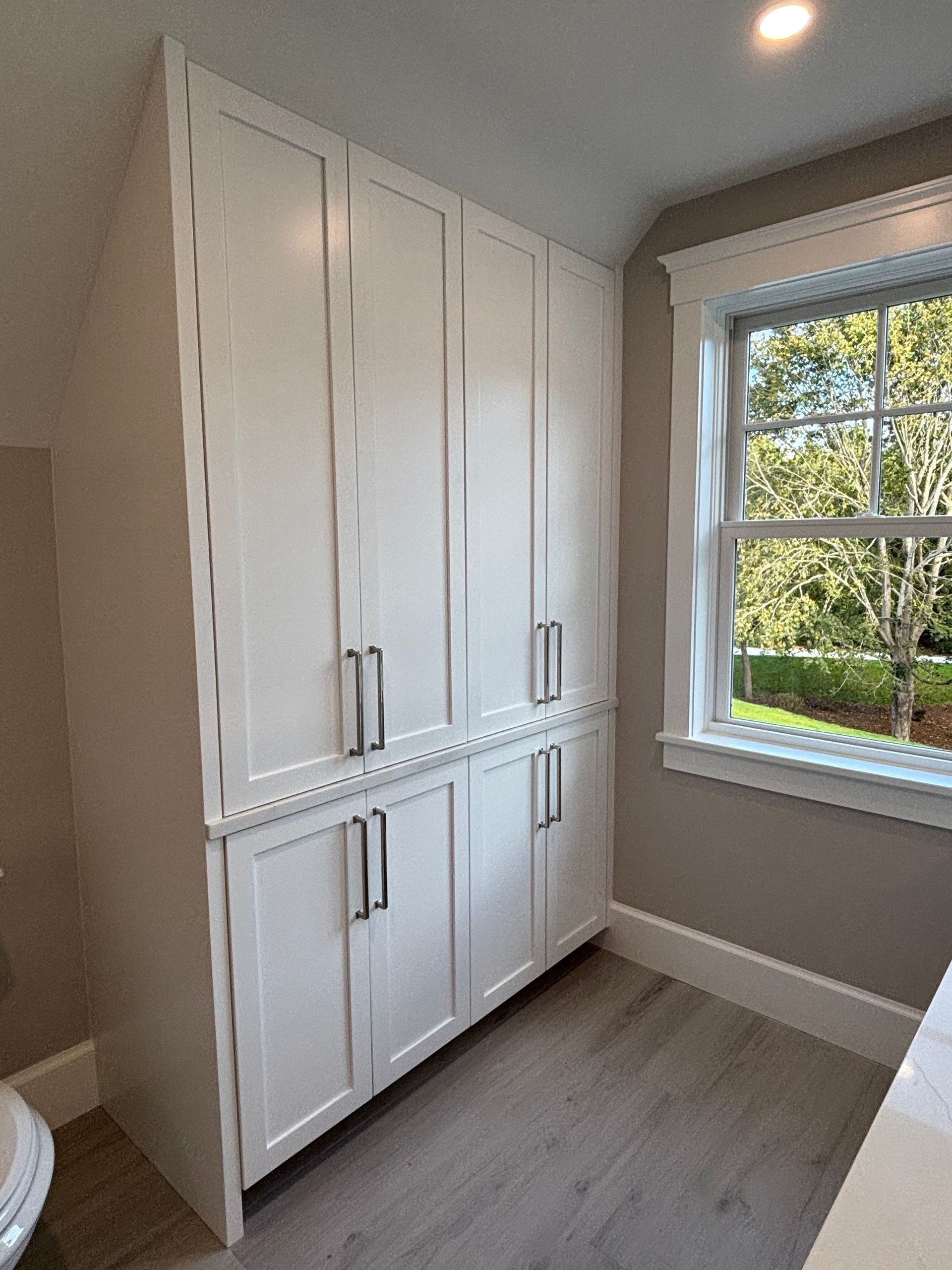 Tall white built-in cabinets with silver handles installed against a light-colored wall next to a window in a room.