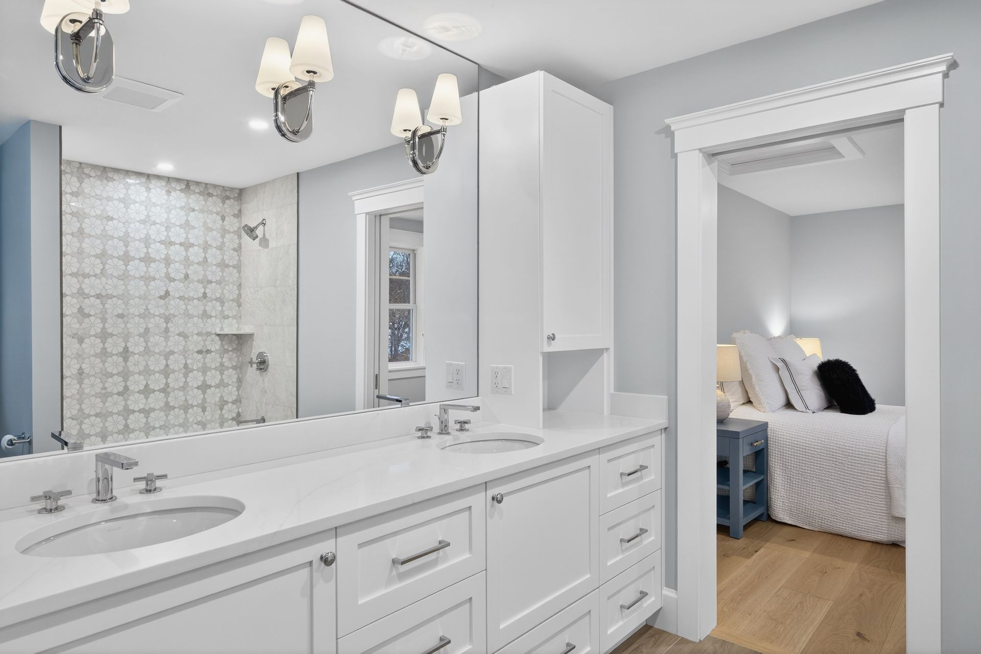 Bathroom with white cabinetry, large mirror, and open doorway to a bedroom with a bed.