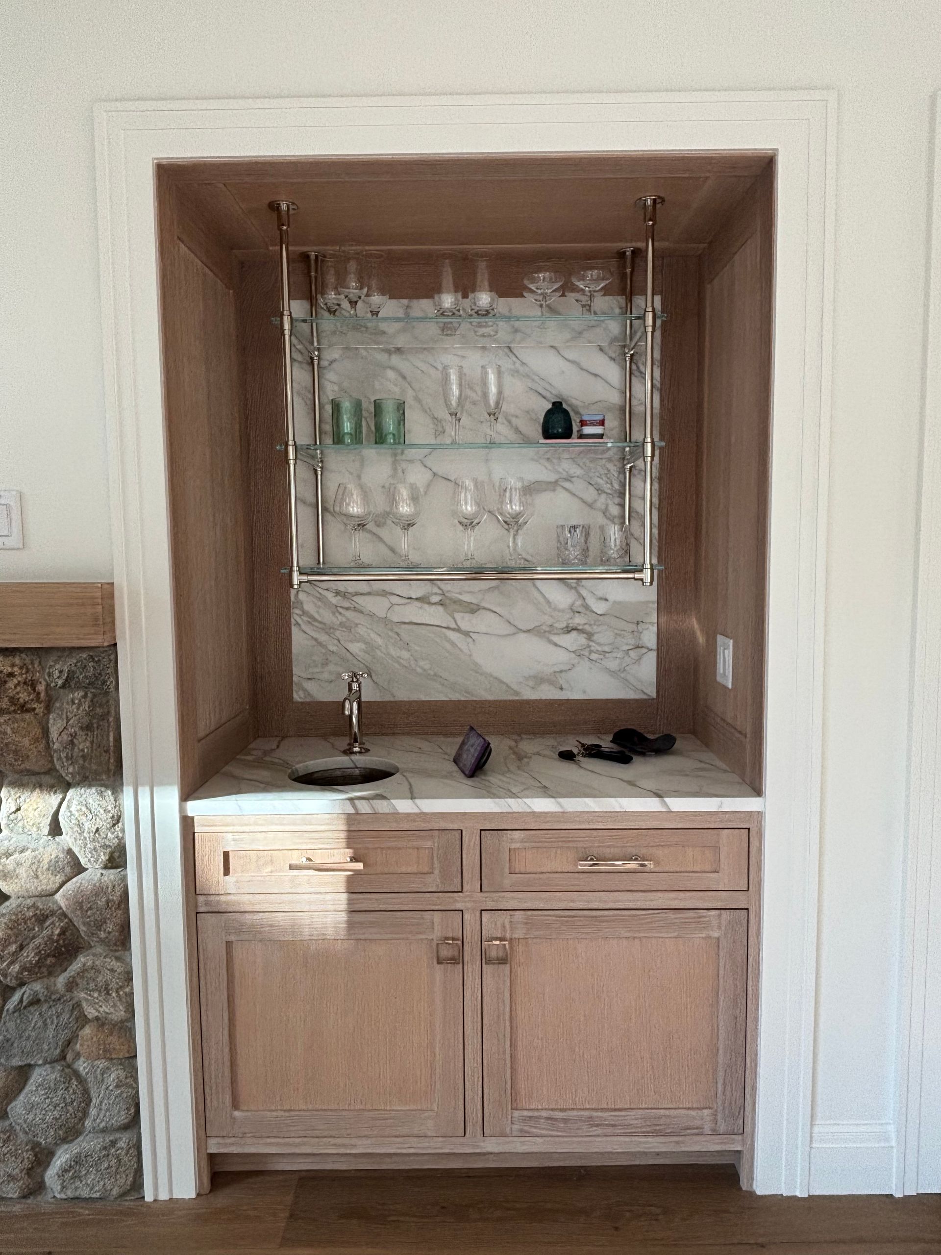 A built-in wet bar with wooden cabinetry, a marble countertop and backsplash, and two glass shelves with glassware.