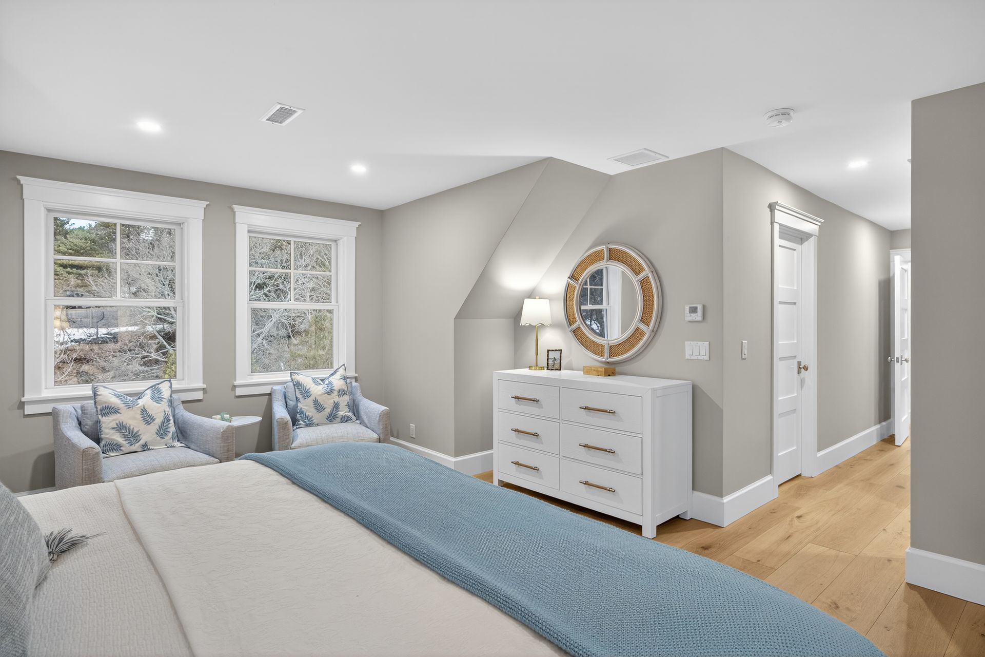 Bedroom with two blue chairs, white dresser, large windows, blue bedspread, and wooden floor.