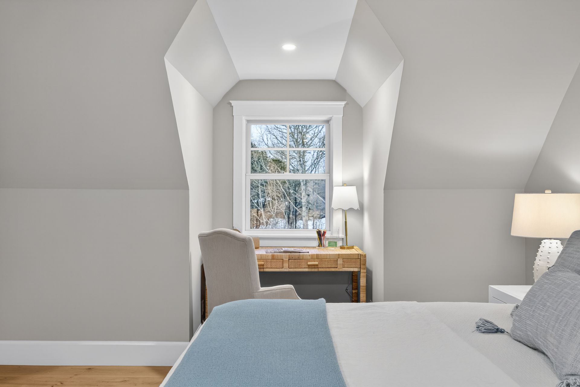 Bedroom with sloped ceiling, desk under window, white walls, and bed with blue blanket.