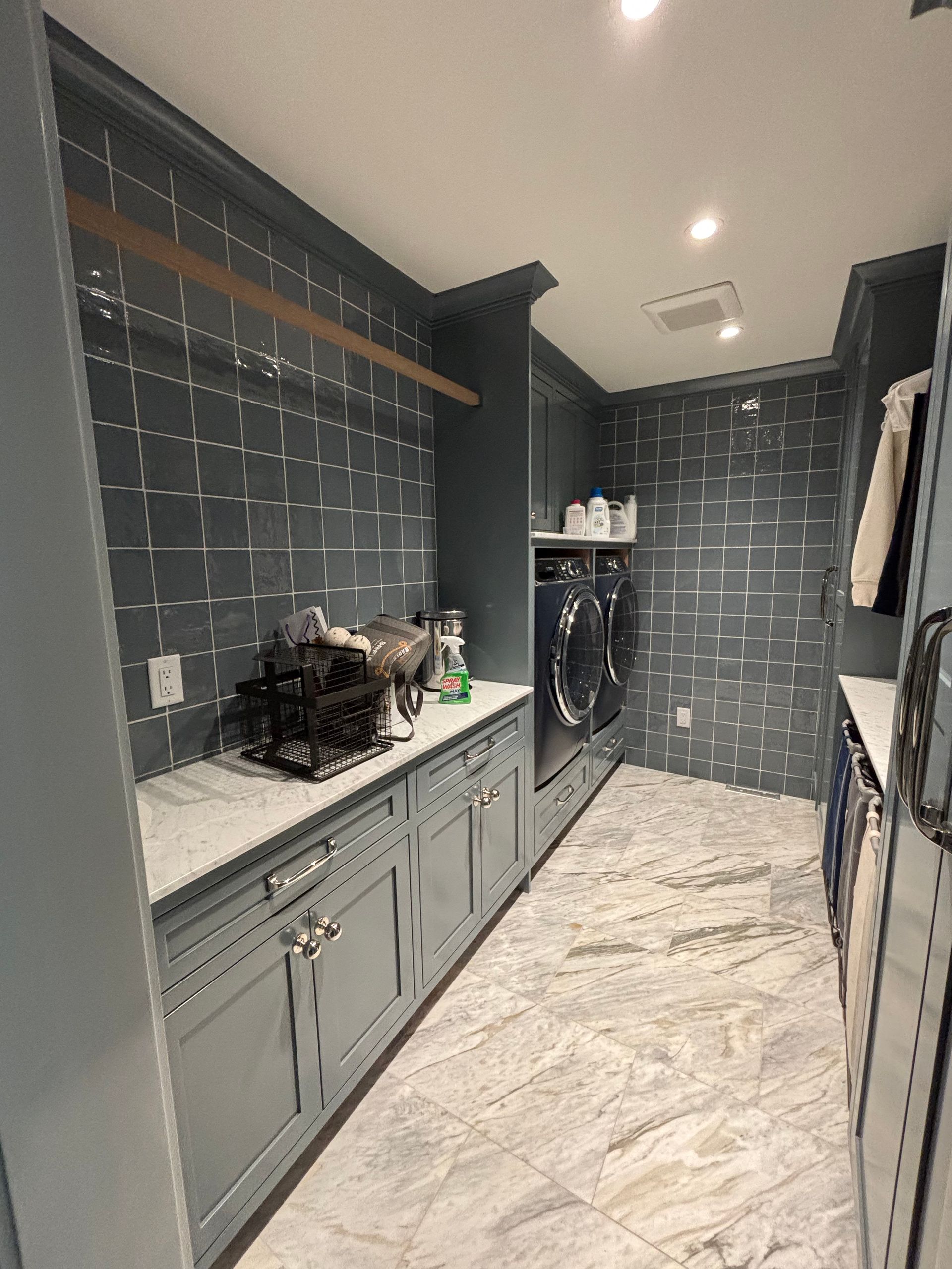 A laundry room with blue-grey walls, patterned tile, marble flooring, grey cabinets, and a washer and dryer unit.