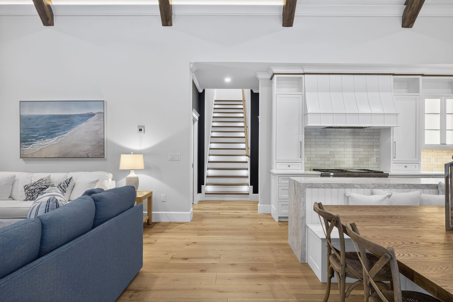 Open-concept living space with a blue sofa, kitchen, staircase, and wooden beams.