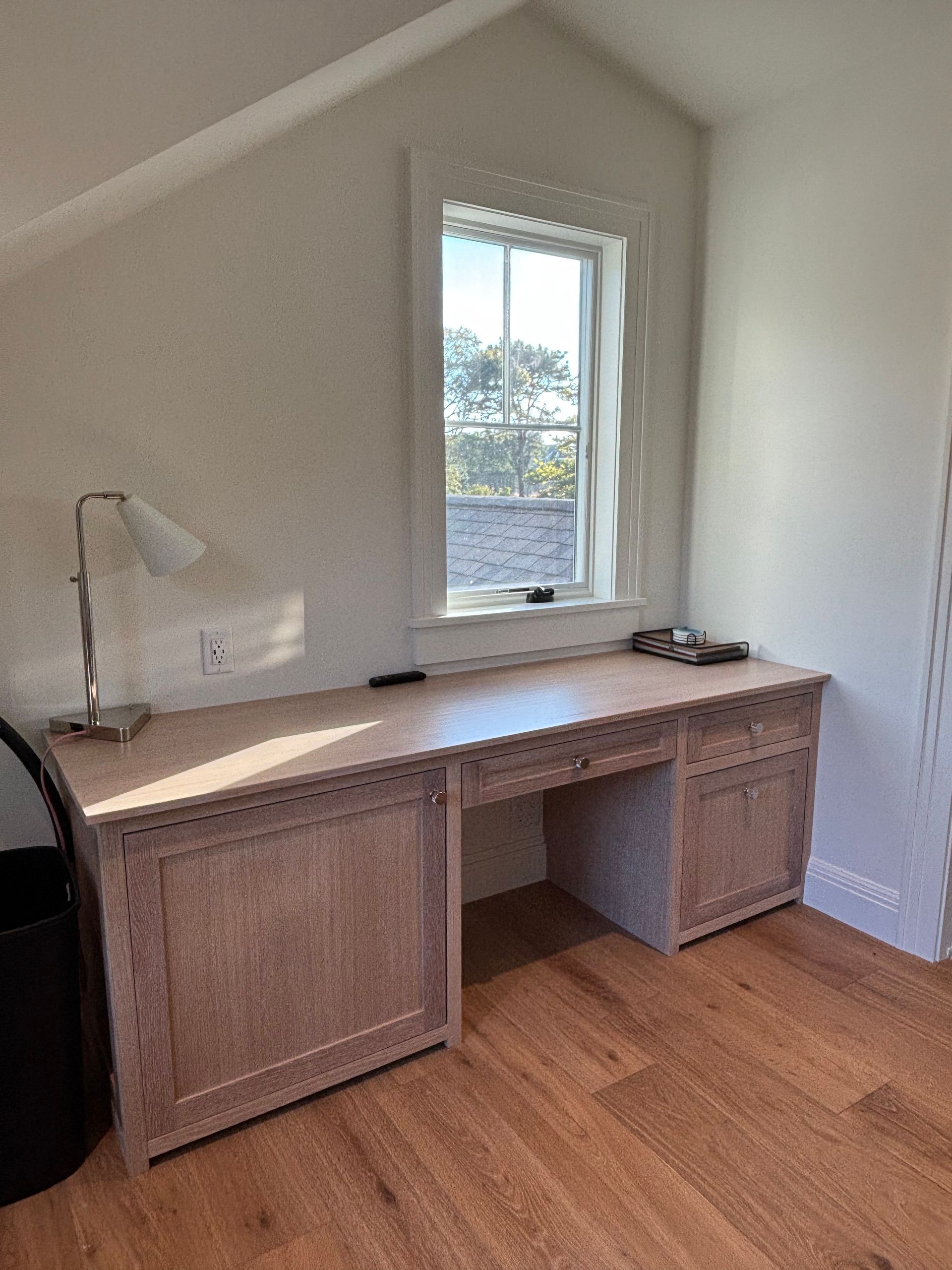 A light-wood built-in desk sits beneath a window in a room with angled walls and hardwood flooring.