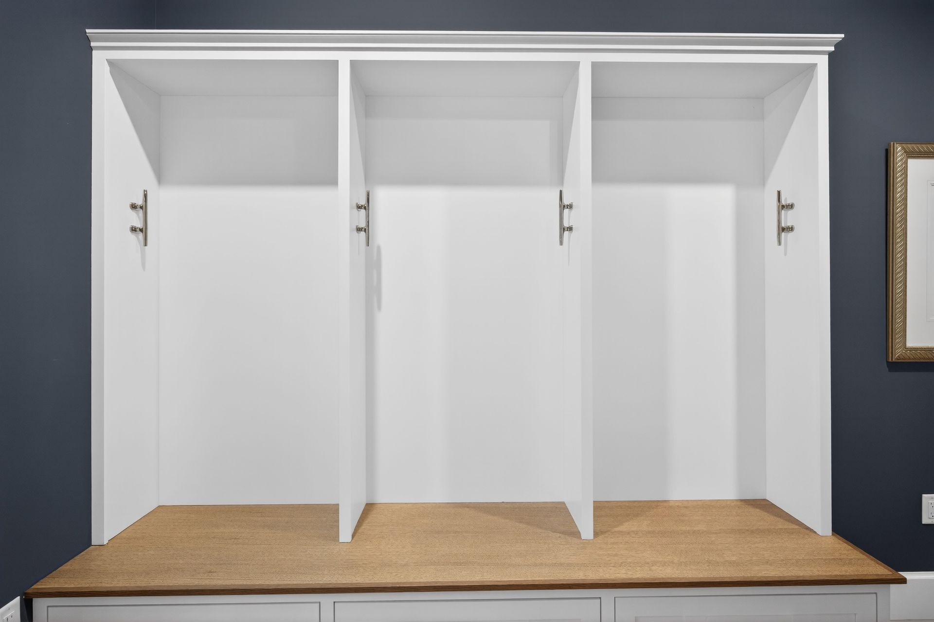 White built-in storage unit with three open sections, brass hardware, and a tan seat. Dark blue wall.