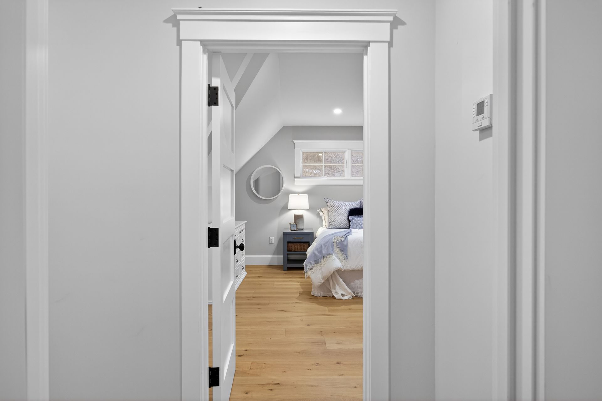 Open doorway into a bedroom with a bed, nightstand, and mirror; light wood floors, white walls.