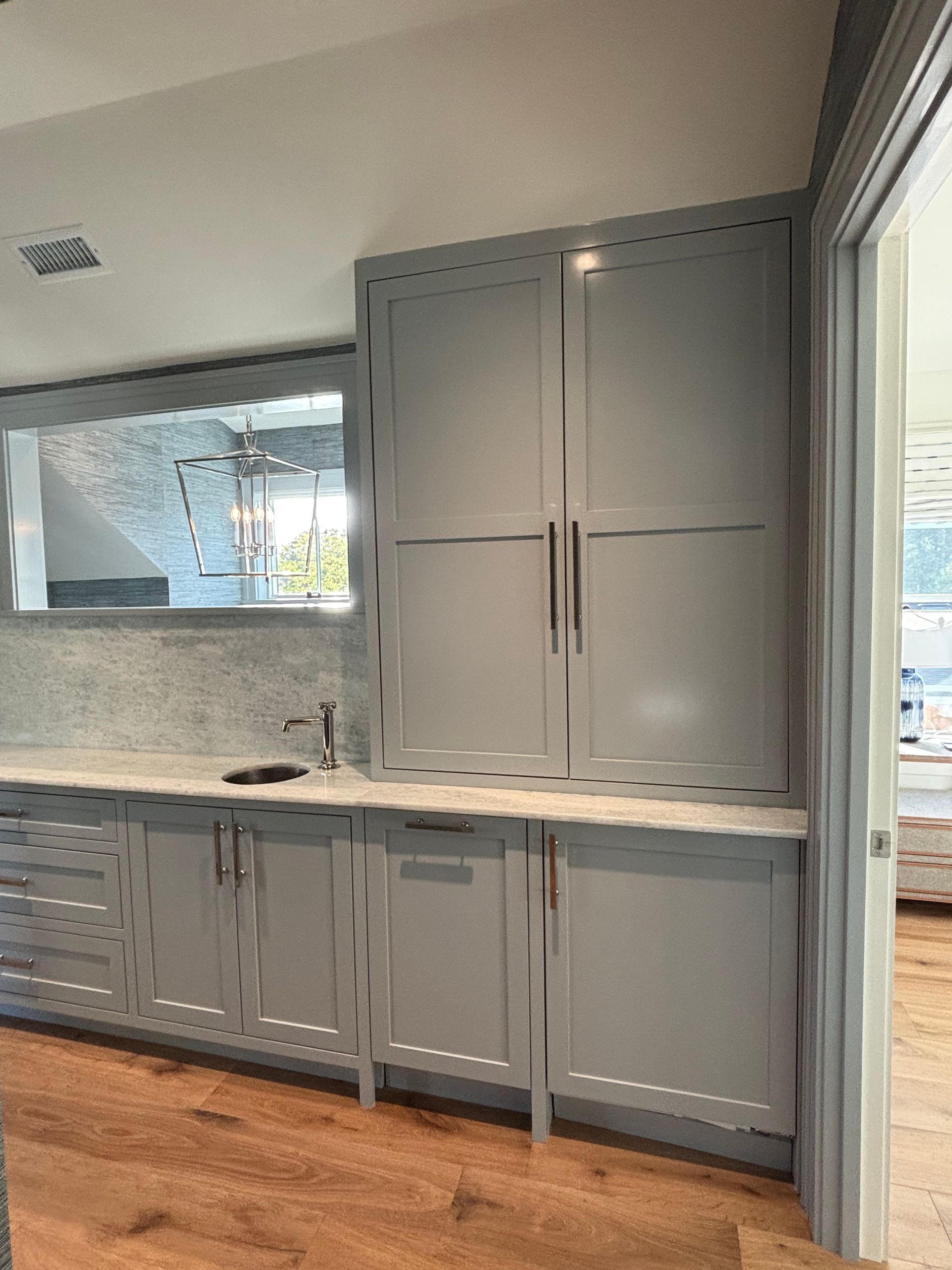 Light gray kitchen cabinets with tall pantry units, a white countertop, and a mirrored backsplash on wood flooring.