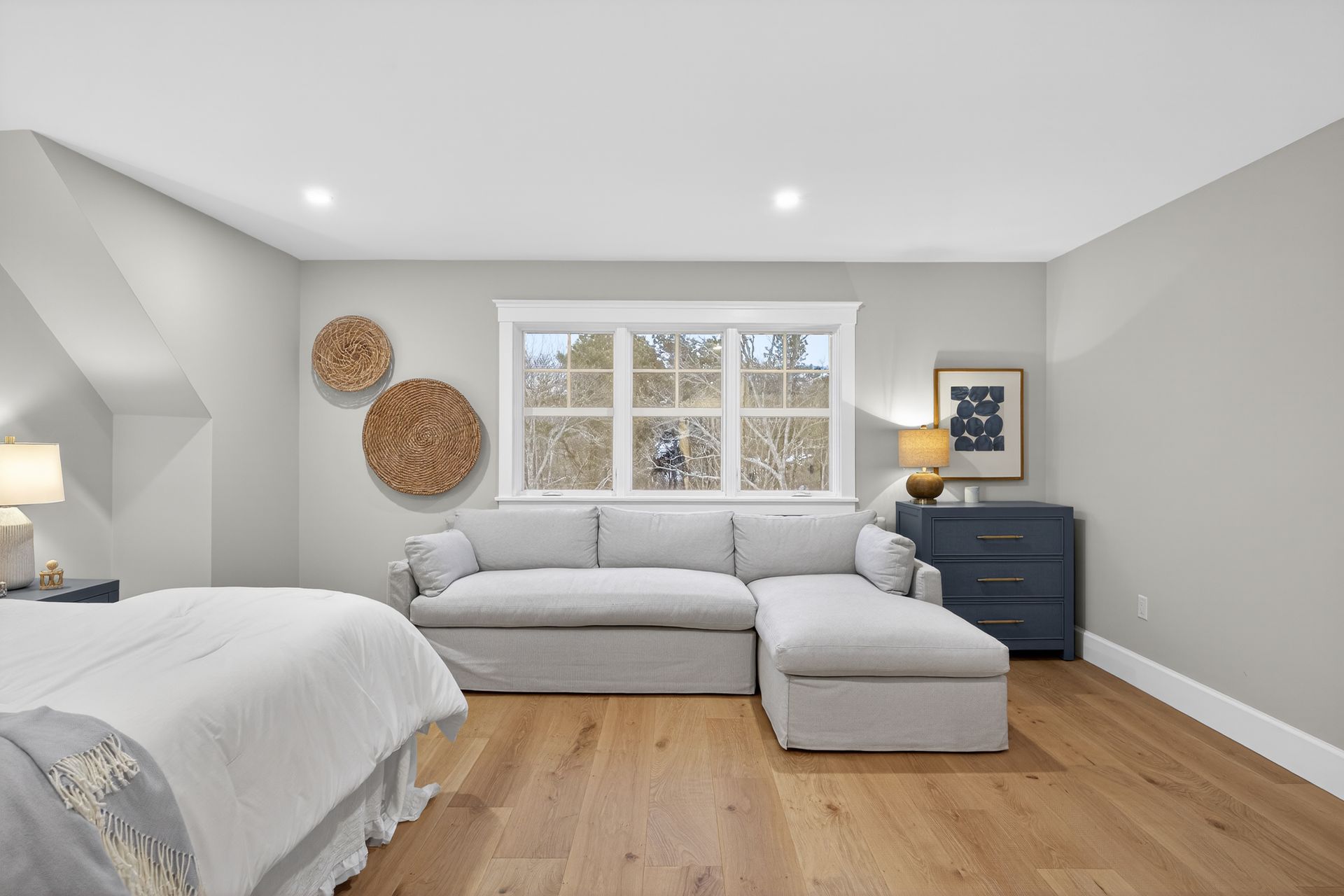 Bedroom with gray sectional sofa in front of a window, hardwood floors, and light gray walls.