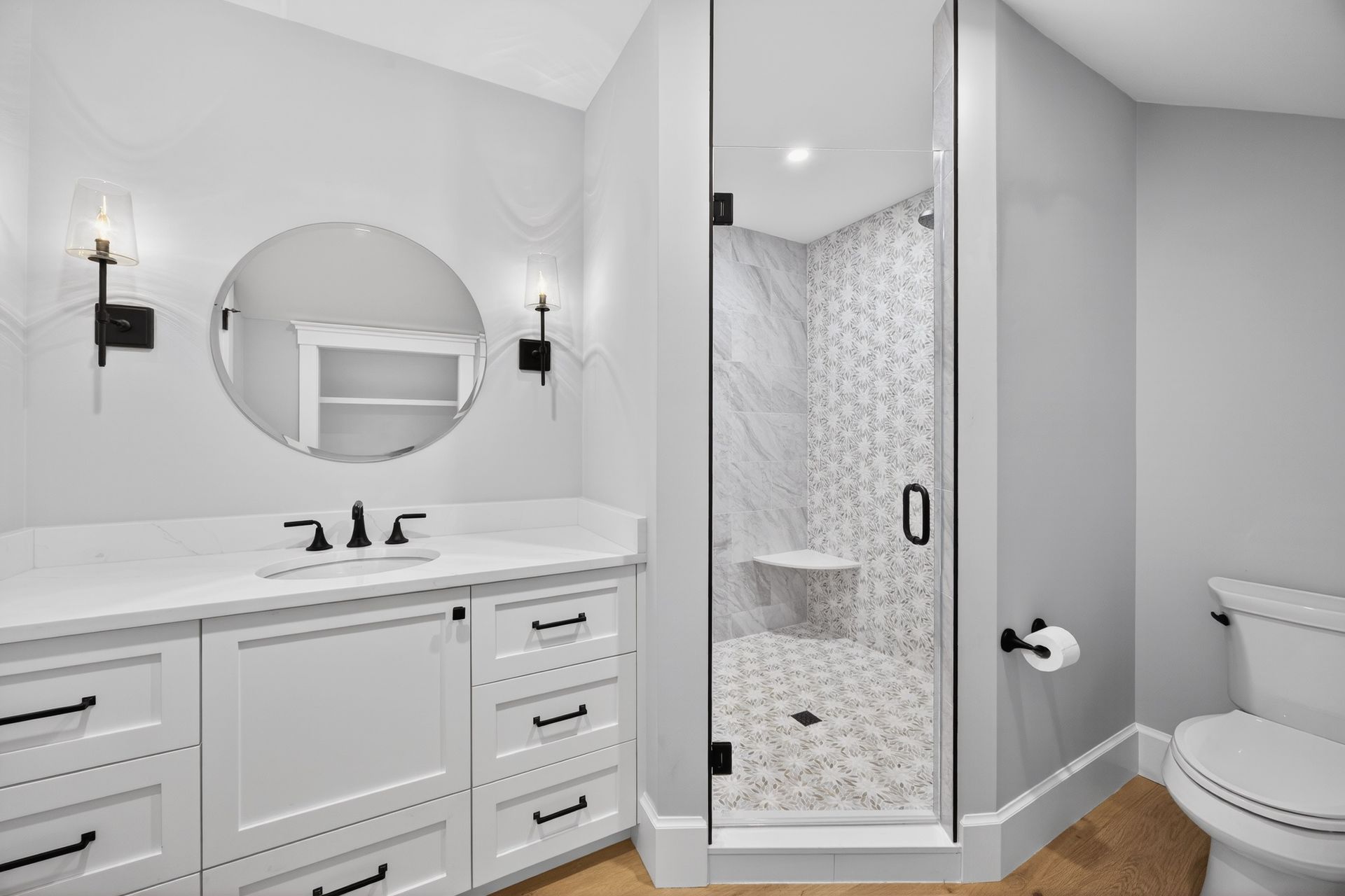 White bathroom with glass shower, white vanity, round mirror, and toilet.