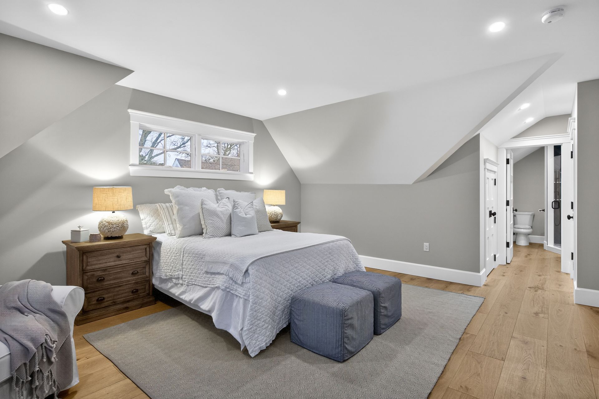 Bedroom with a bed, nightstands, and a view into a bathroom. Light gray walls, hardwood floors, and overhead lighting.