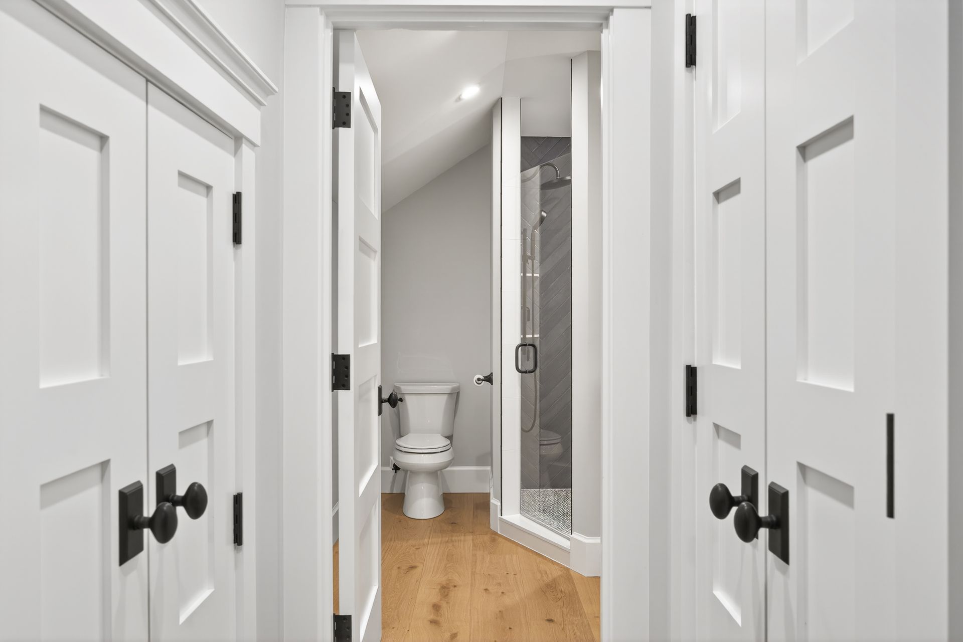 White doors open to a small bathroom with a toilet and shower, light wood floor.