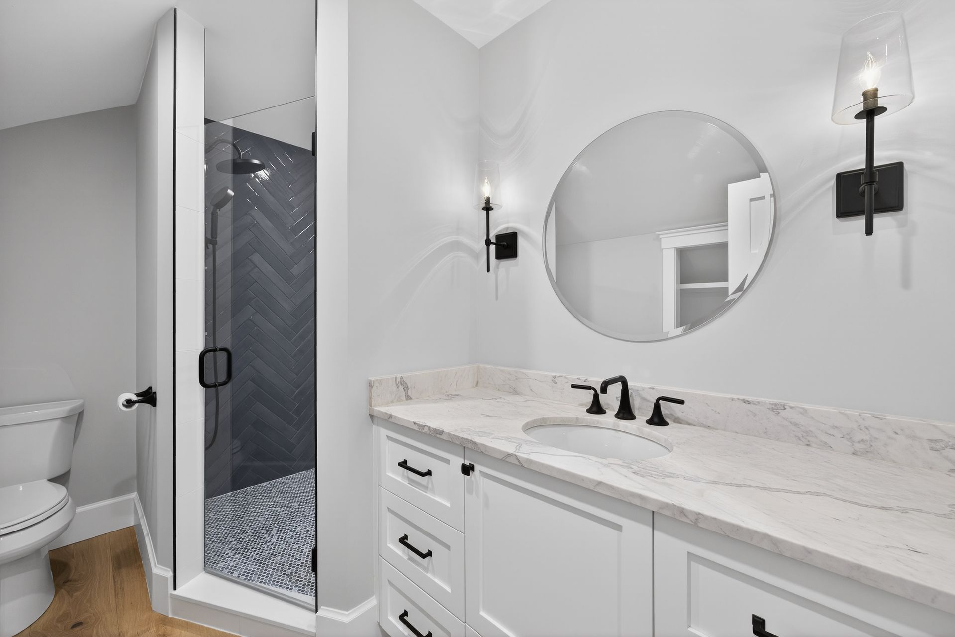 Bathroom with white vanity, round mirror, glass shower with dark tile, and gray walls.