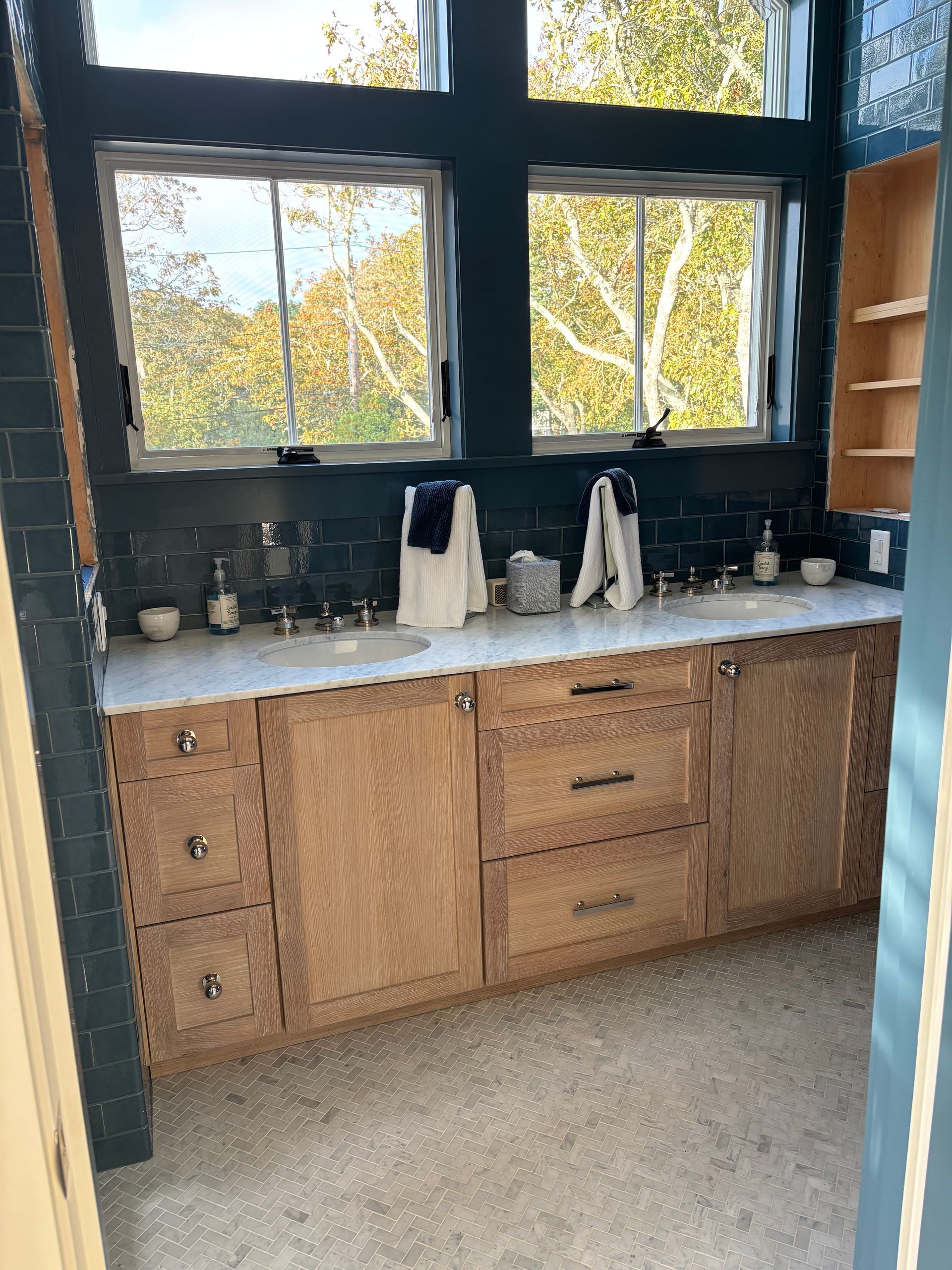 Bathroom vanity with light wood cabinets, white countertops, black tile backsplash, and two windows over the sinks.