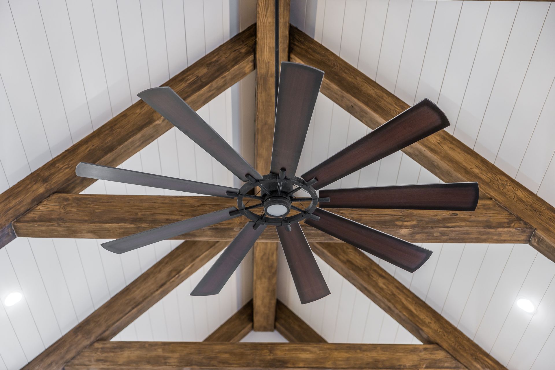 Ceiling fan with multiple dark wooden blades, mounted on wooden beams against a white paneled ceiling.