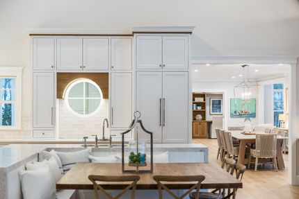 A bright, modern kitchen and dining area featuring white cabinets, a circular window, and a wooden dining table with chairs.