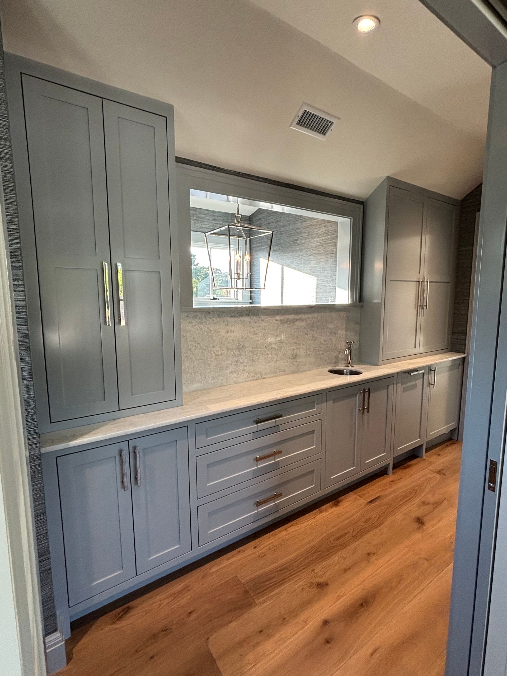 A row of grey cabinets with gold hardware, a marble countertop, and a built-in mirror in a room with hardwood floors.