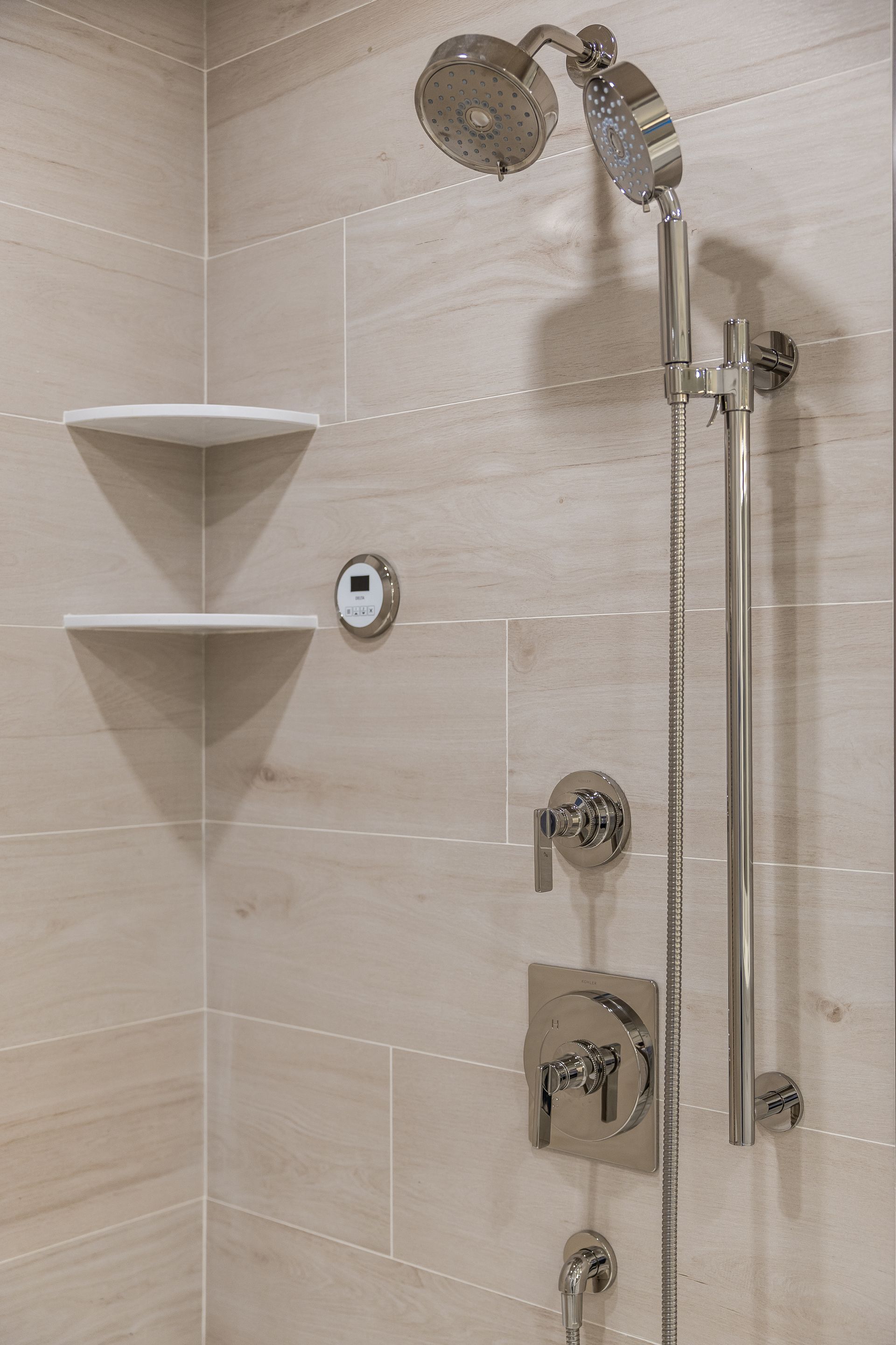 Shower with chrome fixtures, beige tile, two white corner shelves.