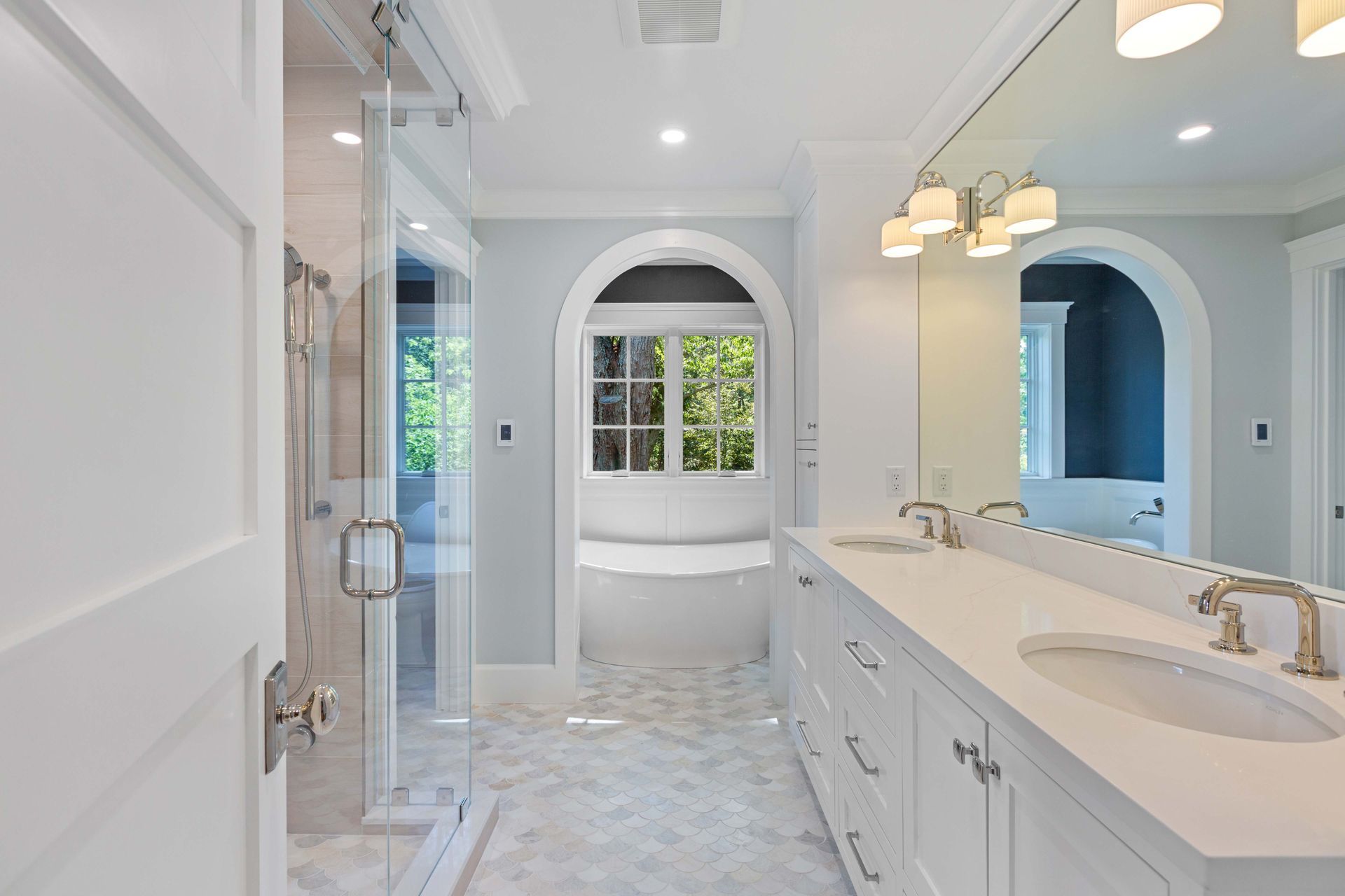 A bright bathroom with a glass shower, double vanity, and a tub viewed through an arched doorway.