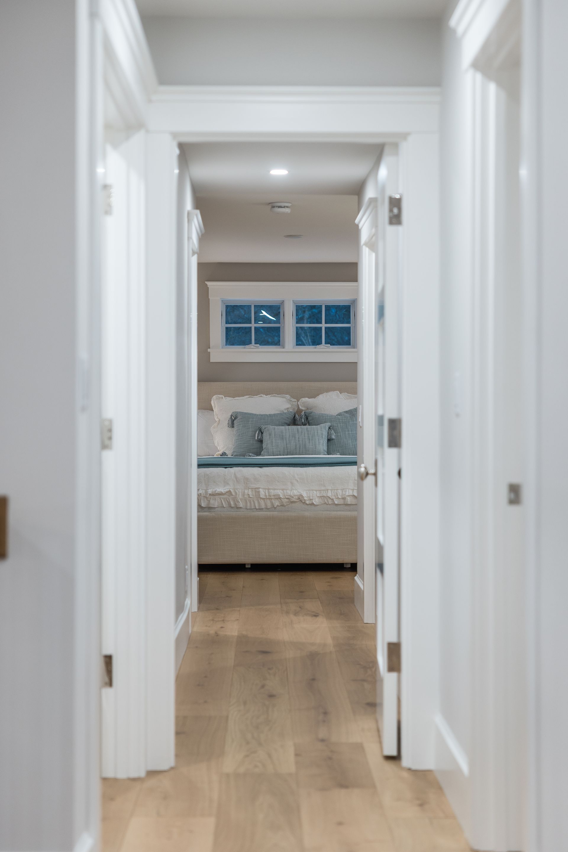 Hallway with light wood floor, leading to a bedroom with light blue bed and small windows.