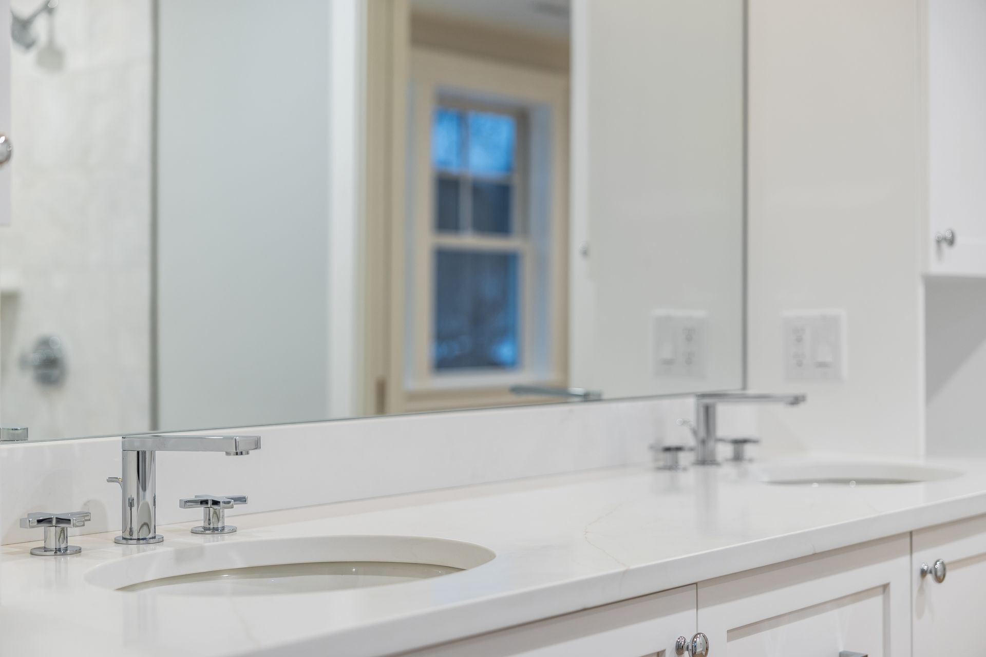 Bathroom with white countertop, two sinks, chrome faucets, and a large mirror reflecting a window.