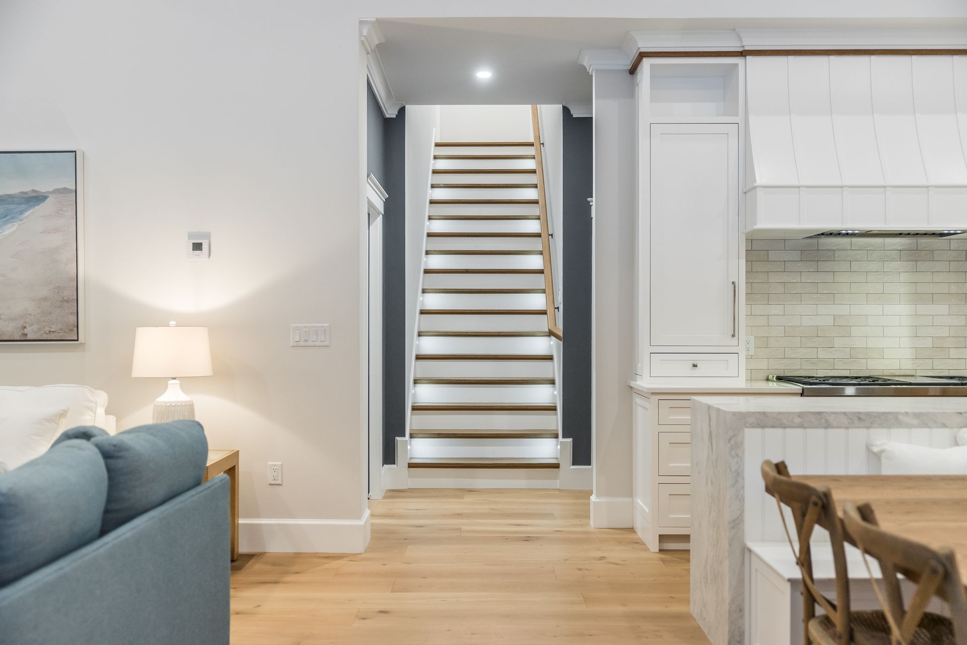 Interior view with staircase, kitchen, and living area. White cabinets, light wood floors, blue sofa.