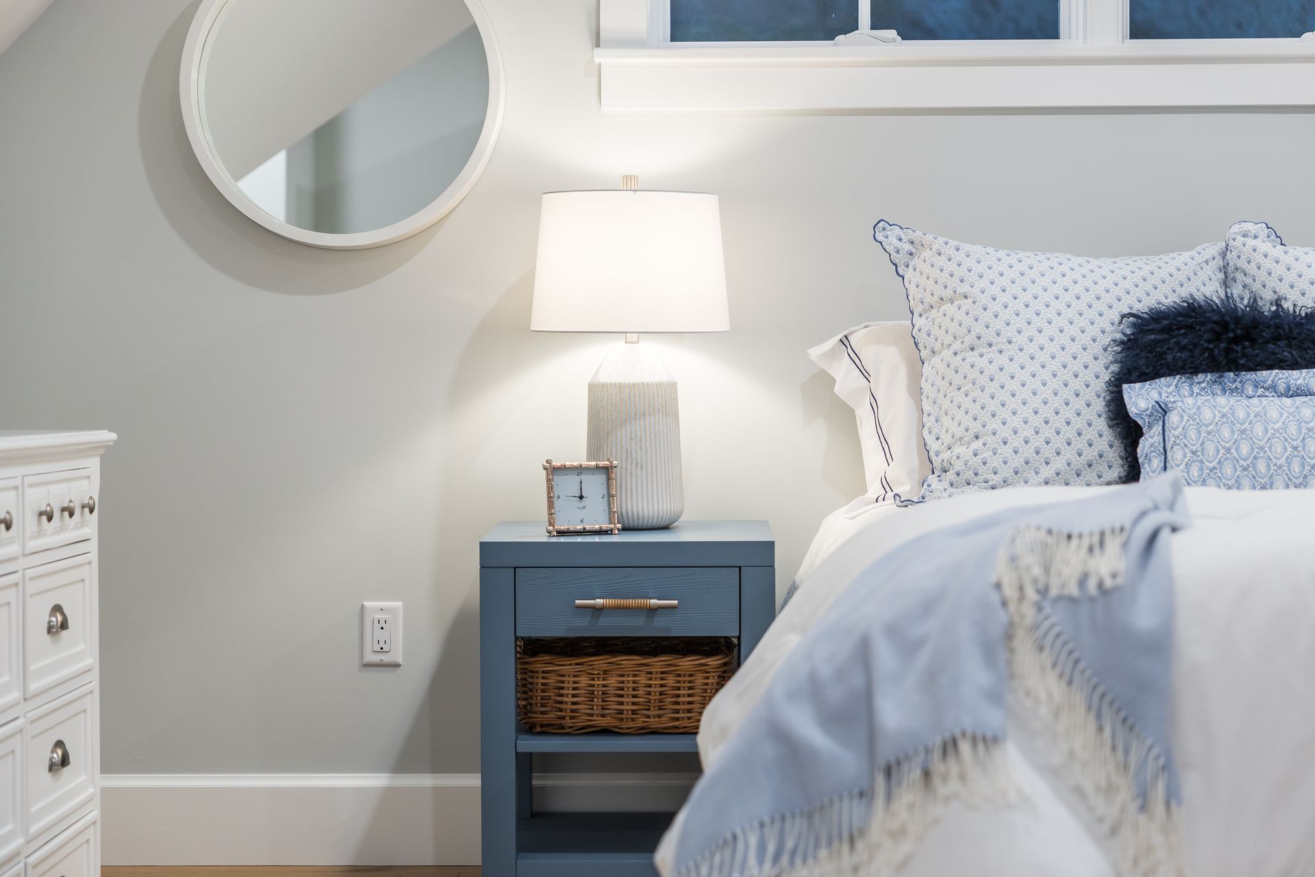 Bedroom scene: blue nightstand with lamp, bed with blue accents, round mirror, white dresser.
