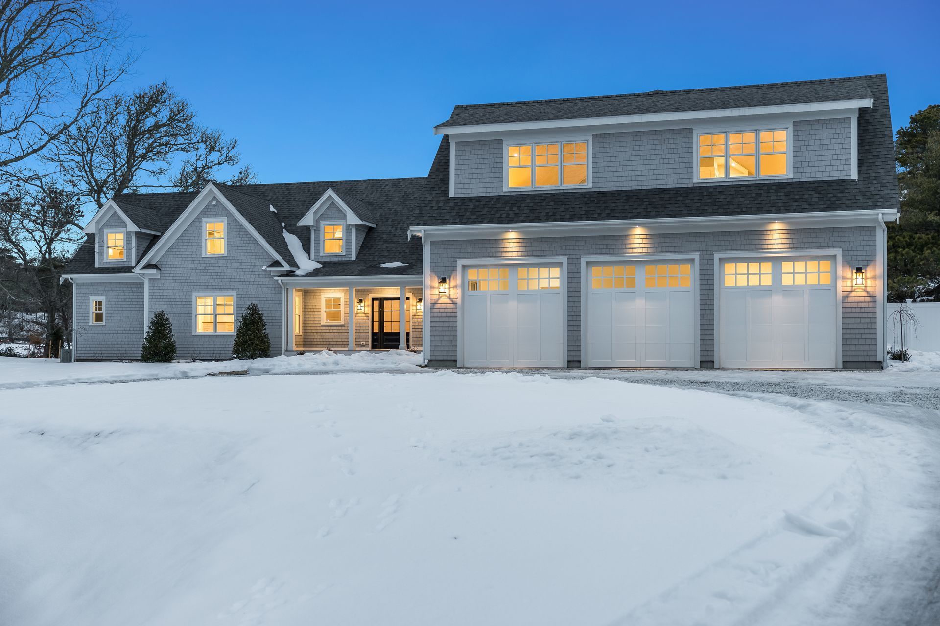 A grey suburban house with a three-car garage, set against a twilight sky with snow covering the front lawn.