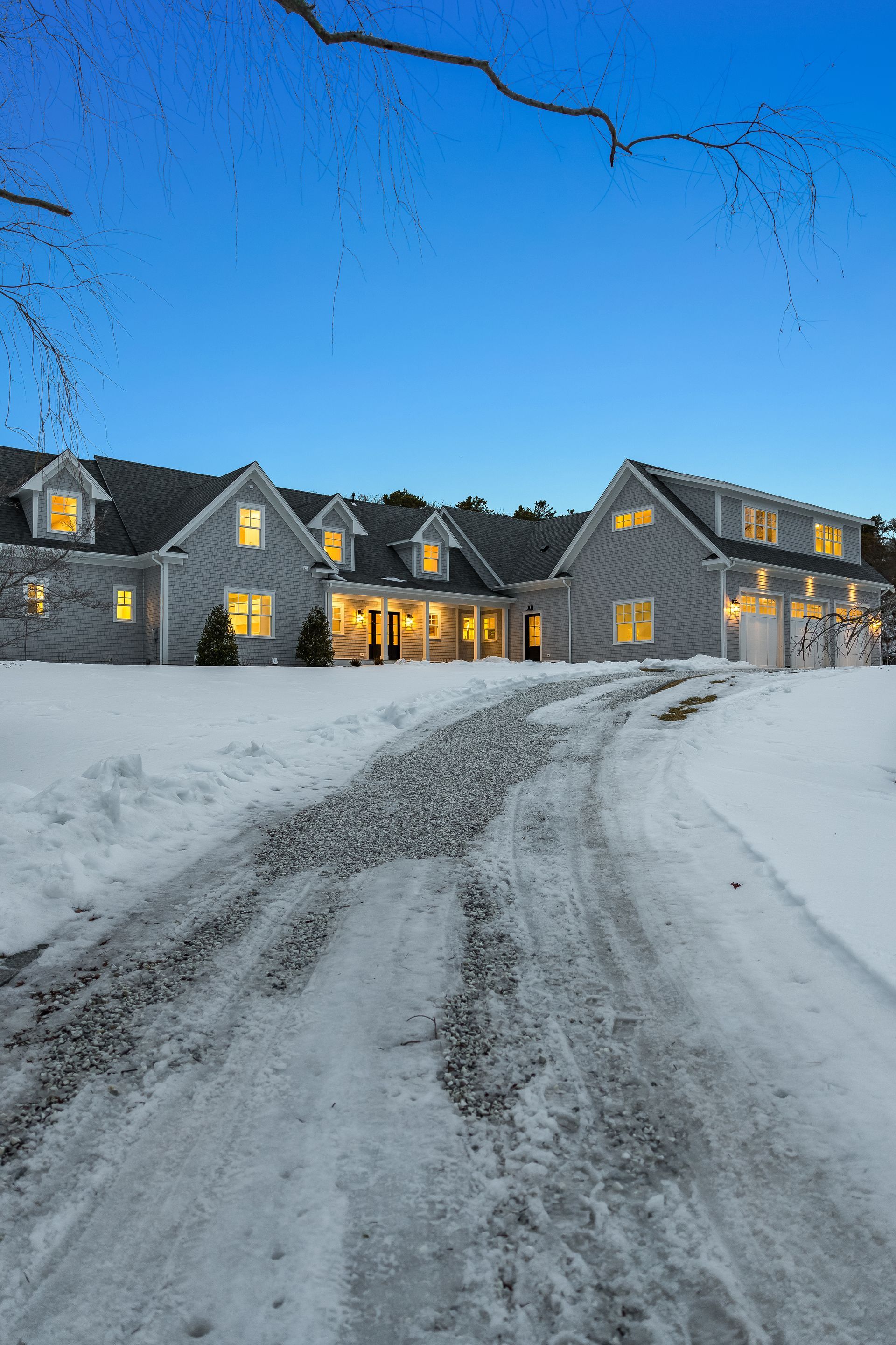 A light-gray multi-story home with glowing warm lights at dusk, surrounded by deep, snow-covered ground and a driveway.